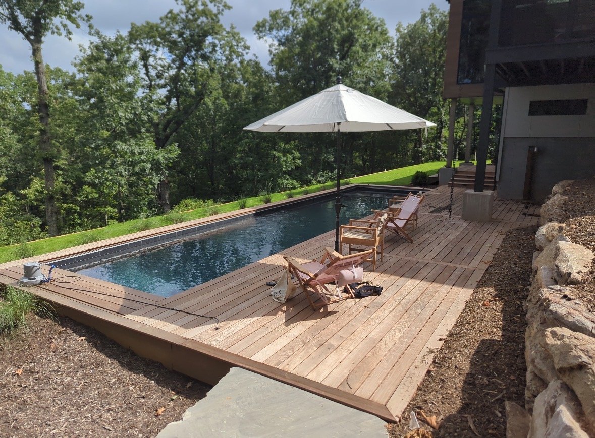 Backyard pool with wooden deck, surrounded by trees, with a large white umbrella and four chairs on the deck.