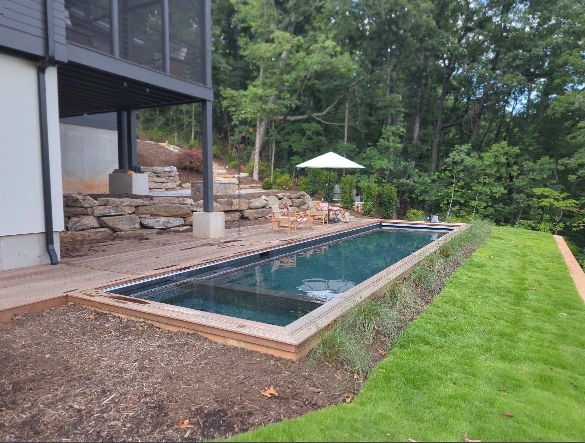 A backyard with a rectangular in-ground swimming pool, a wooden pool deck with outdoor seating, a white patio umbrella, a lush green lawn, and dense trees in the background.