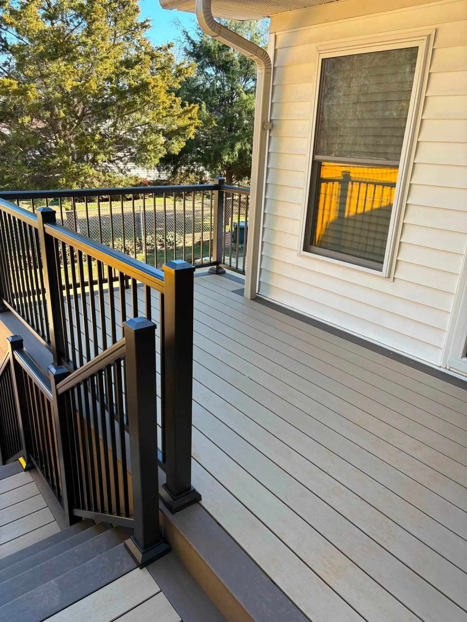 View of a residential balcony with beige wooden flooring, black metal and wooden railing, beige house siding, and a window with closed blinds. In the background, there are green trees and a clear blue sky.