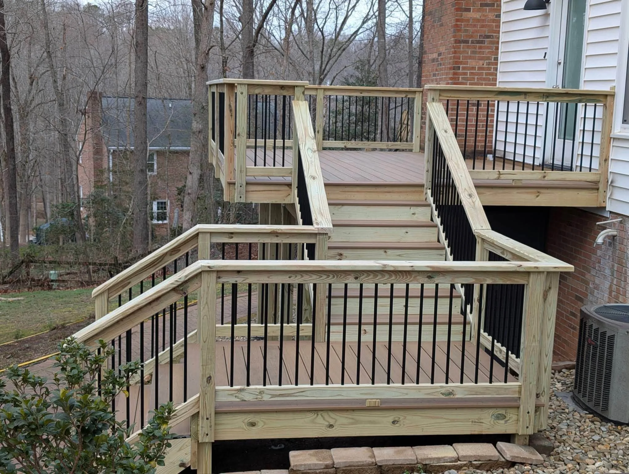 Wooden deck stairs with black metal railing leading up to a backyard deck attached to a house with brick and white siding.