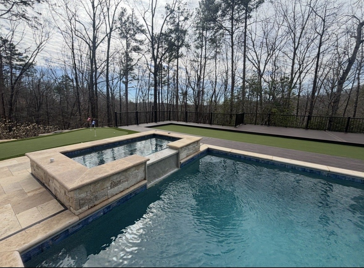 Backyard with swimming pool, hot tub, artificial grass, wooden deck, and a wooded area with bare trees in the background.