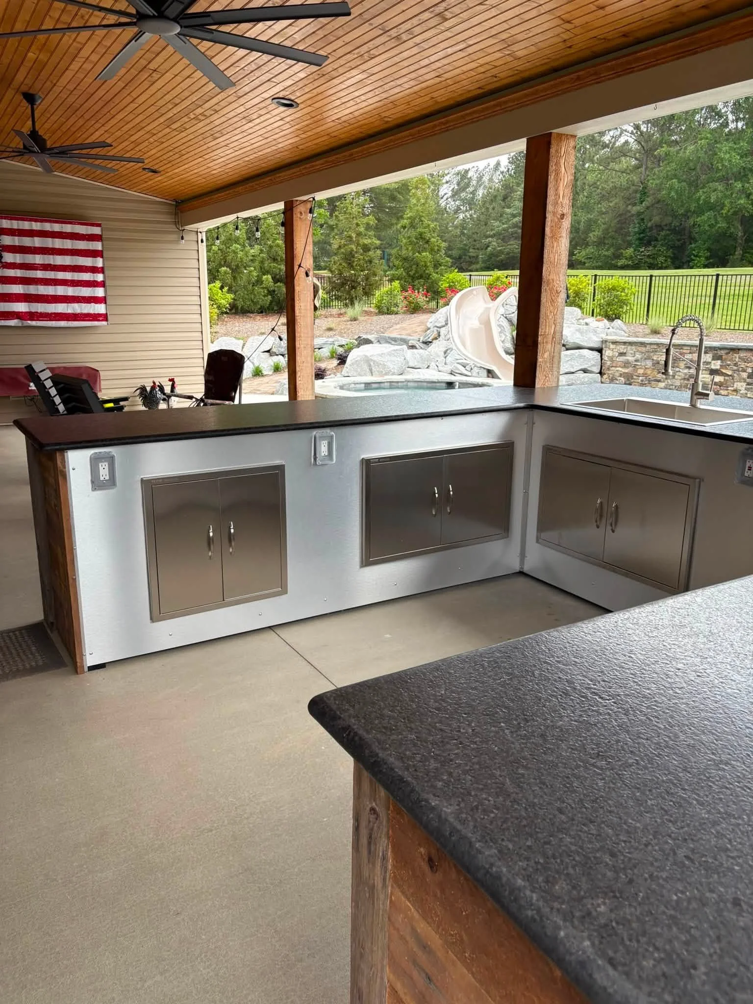 View of an outdoor backyard area with a covered patio, ceiling fans, an American flag wall art, a table with chairs, a small pool with a white slide, surrounded by rocks, plants, and trees, and a black metal fence enclosing the yard.