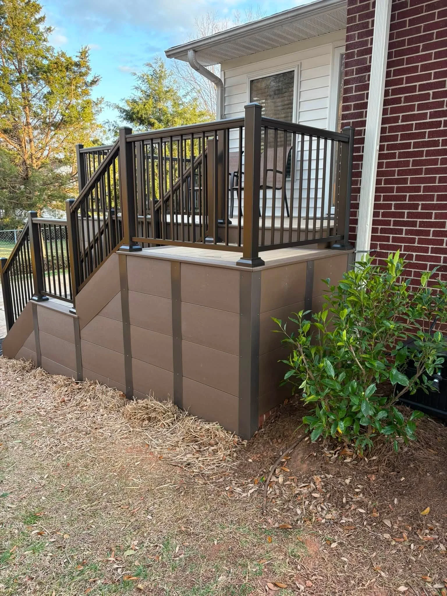 A small deck with stairs leading to a backyard with trees and a chain-link fence. The deck has black metal railing, and there is a brown chair and a small table on it. The house exterior features white siding and red brick wall, and a white downspout