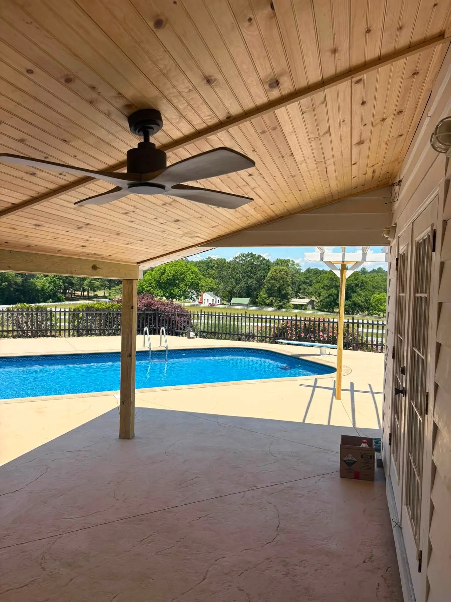 View of a backyard patio with a hanging ceiling fan, a swimming pool, and trees in the background. The patio has a wooden ceiling and concrete flooring.