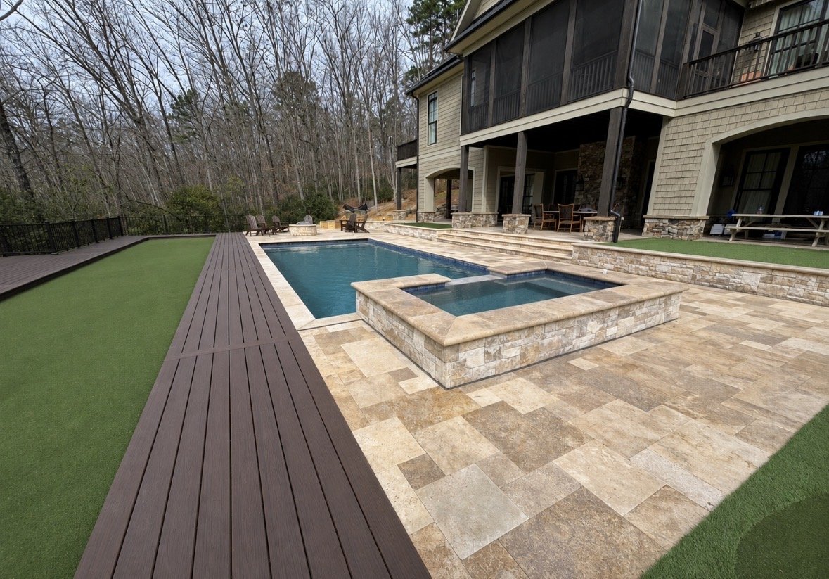 Backyard with a swimming pool and hot tub, wooden deck, stone patio, outdoor furniture, and a house with a balcony, surrounded by trees.