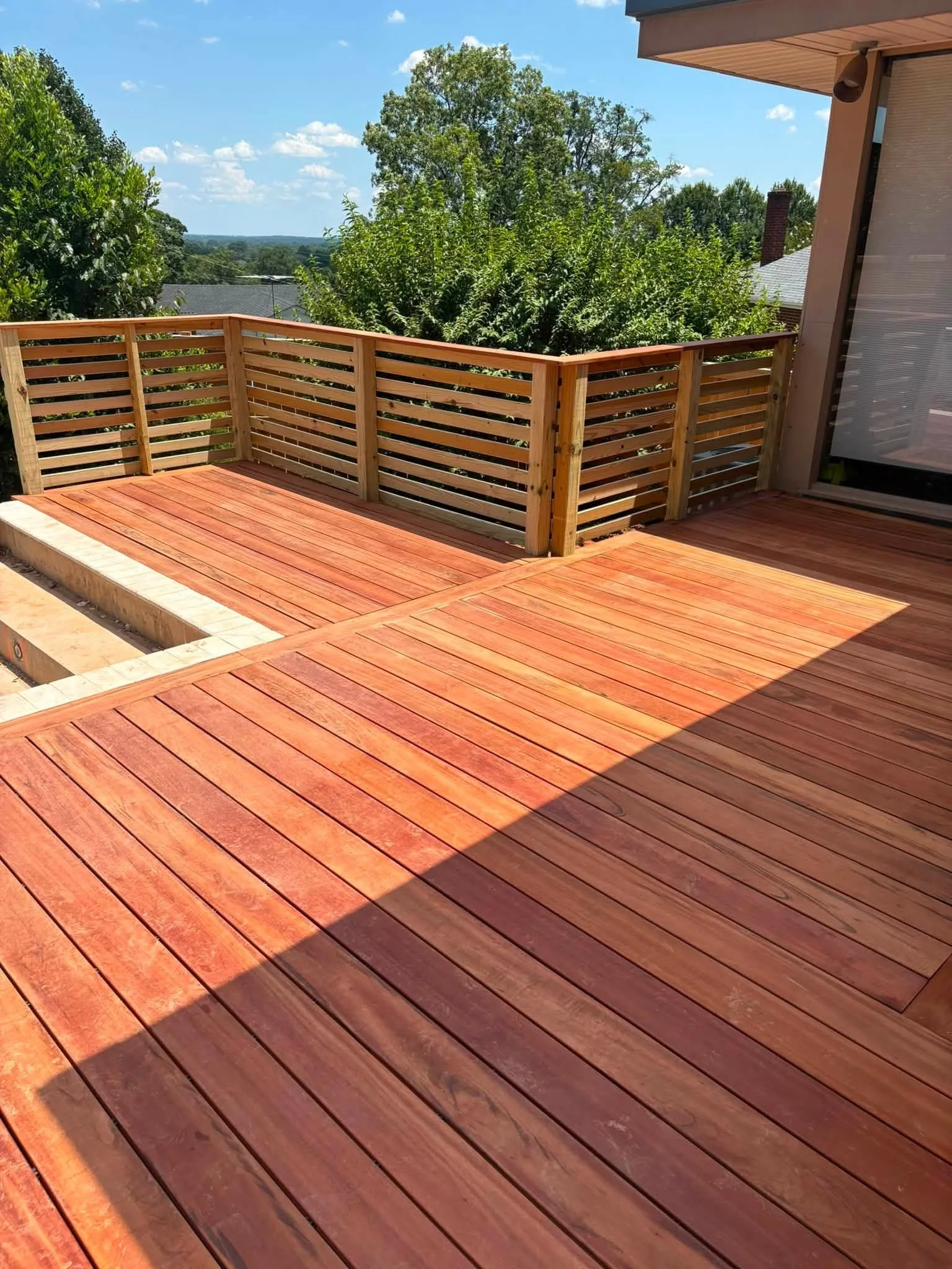 A wooden deck with a corner railing, surrounded by green trees under a clear blue sky with some white clouds.