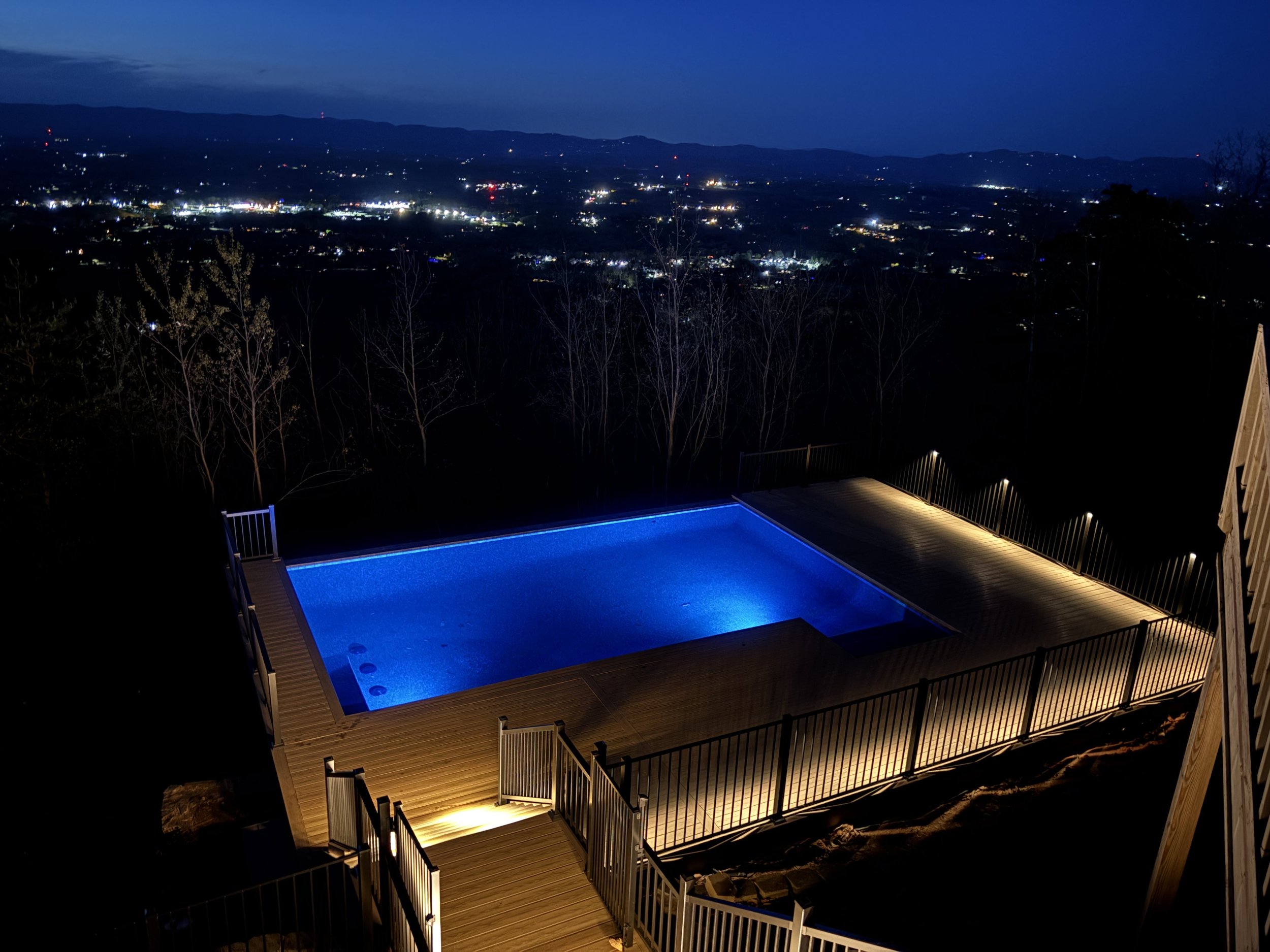 Night view of a city and mountains in the distance, with a wooden deck that has a lit railing and a pool with blue lighting in the foreground.