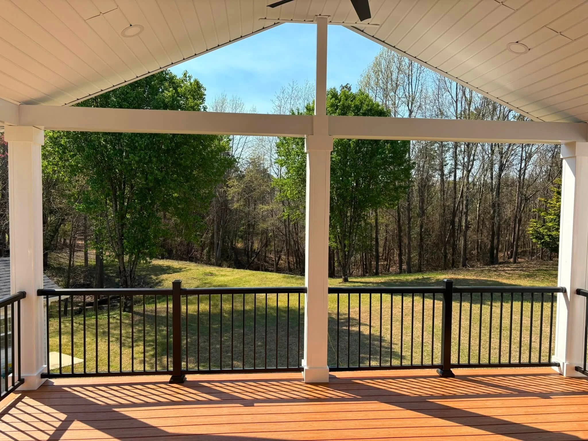 View from a balcony with a wooden floor, black metal railing, and white support columns, overlooking a lush green backyard with trees and grass under a clear blue sky.