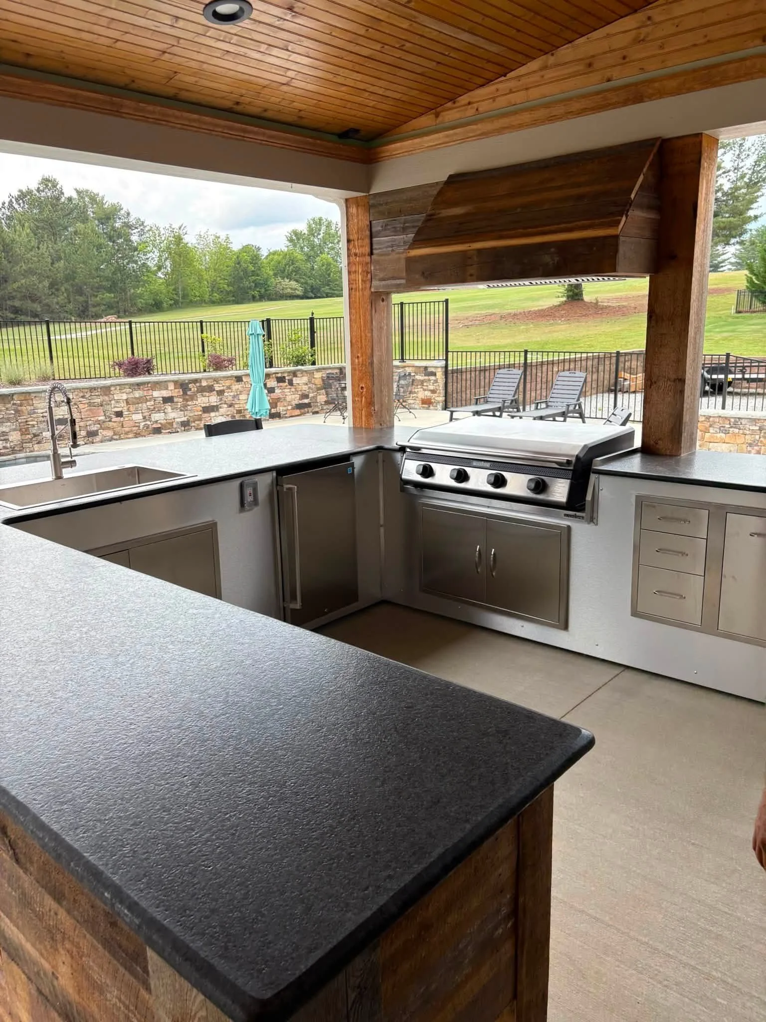 Outdoor kitchen area with a black granite counter, stainless steel appliances, and a scenic backyard with lawn chairs and a fence.