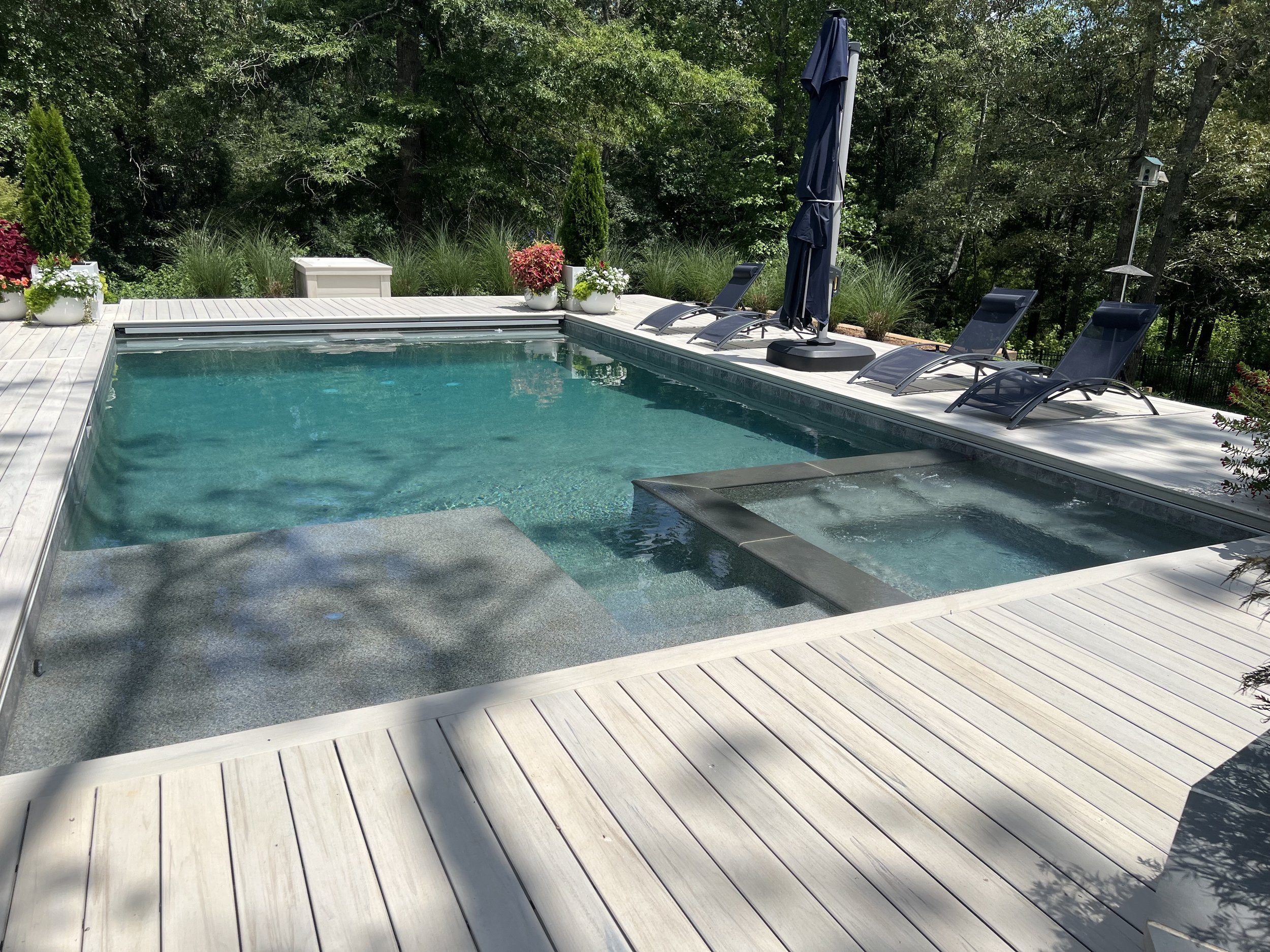 A backyard swimming pool with a wooden deck surrounded by greenery, including trees and plants; several lounge chairs and a closed umbrella are positioned on the deck.