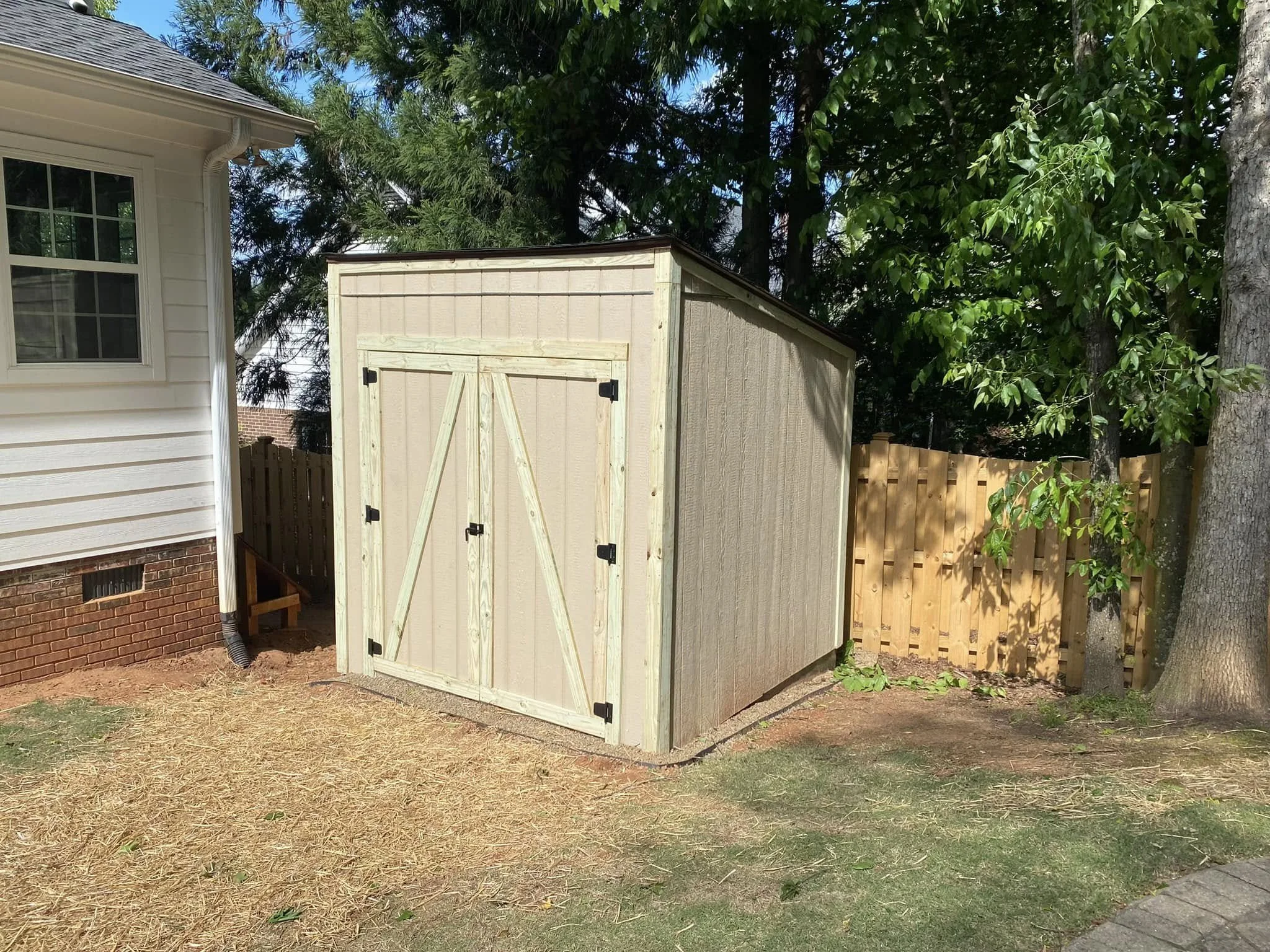 Backyard with a wooden shed, a house wall on the left, trees, a fence, and a barren ground surface.