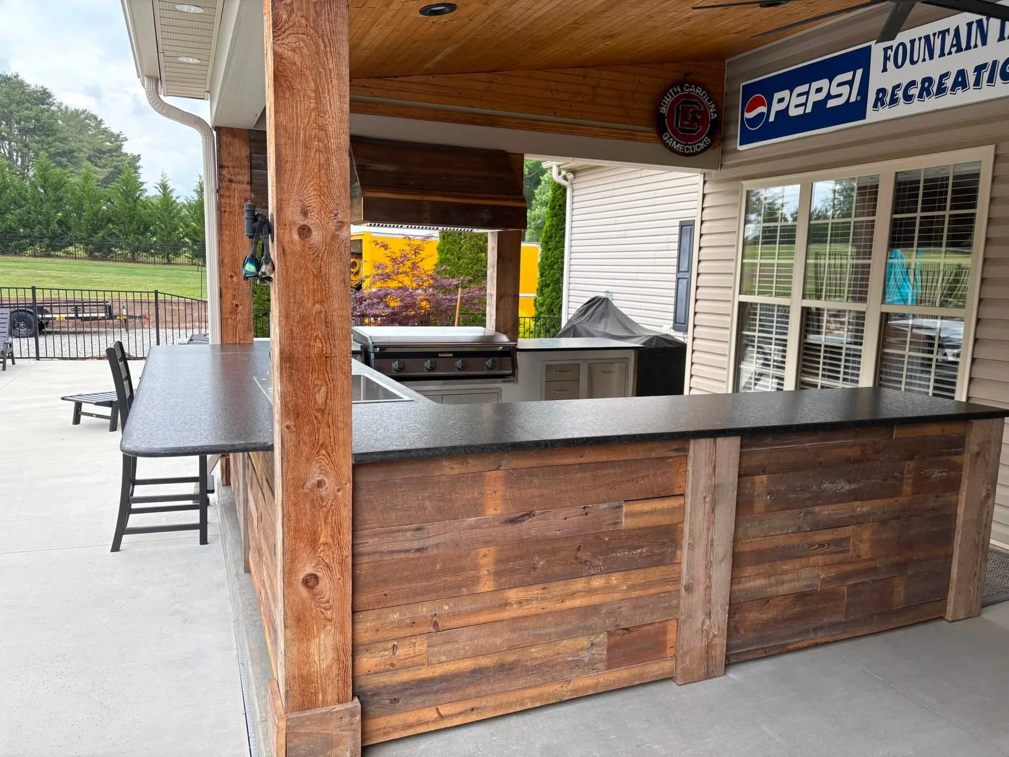 Outdoor kitchen area with a granite countertop, a grill, and a covered wood structure. There are chairs and a fence in the background with trees and a field.
