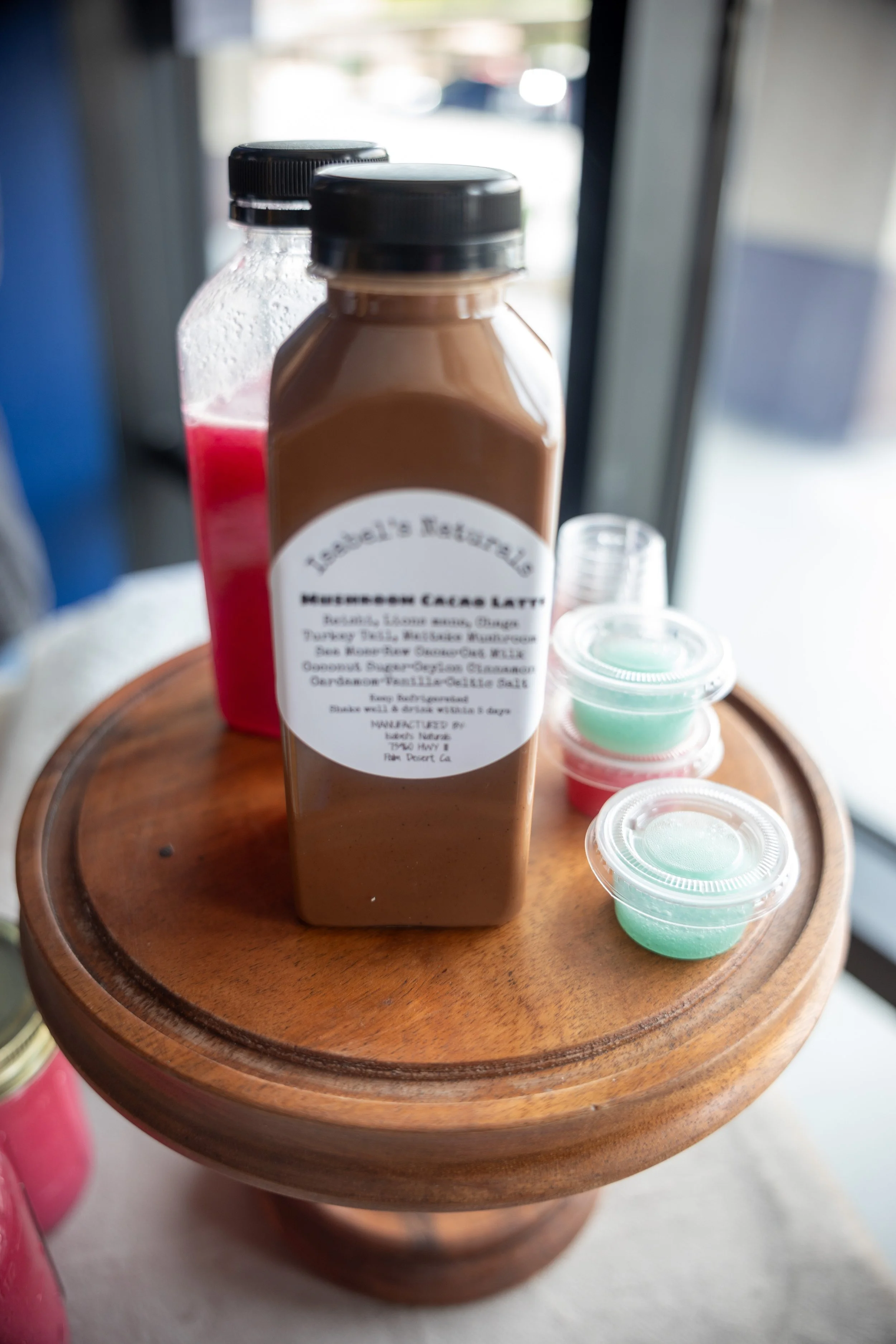 A wooden tray holding a bottle of mocha chocolate latte and small cups of colorful drinks near a window.