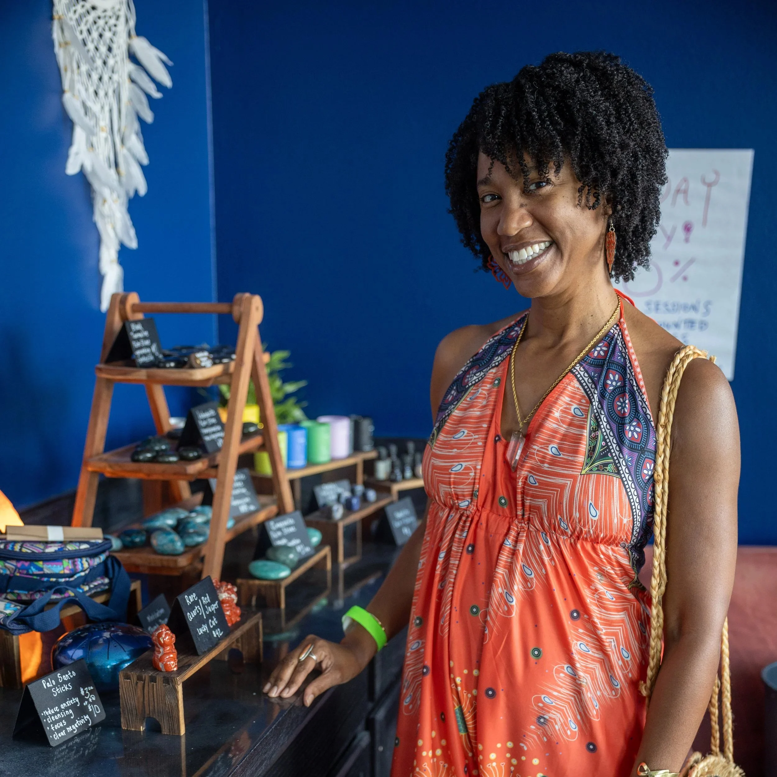 A woman with curly black hair smiling and wearing a colorful dress with orange, blue, and white patterns, standing next to shelves of various small products at a market or shop. The background features a blue wall and a paper with partially visible w