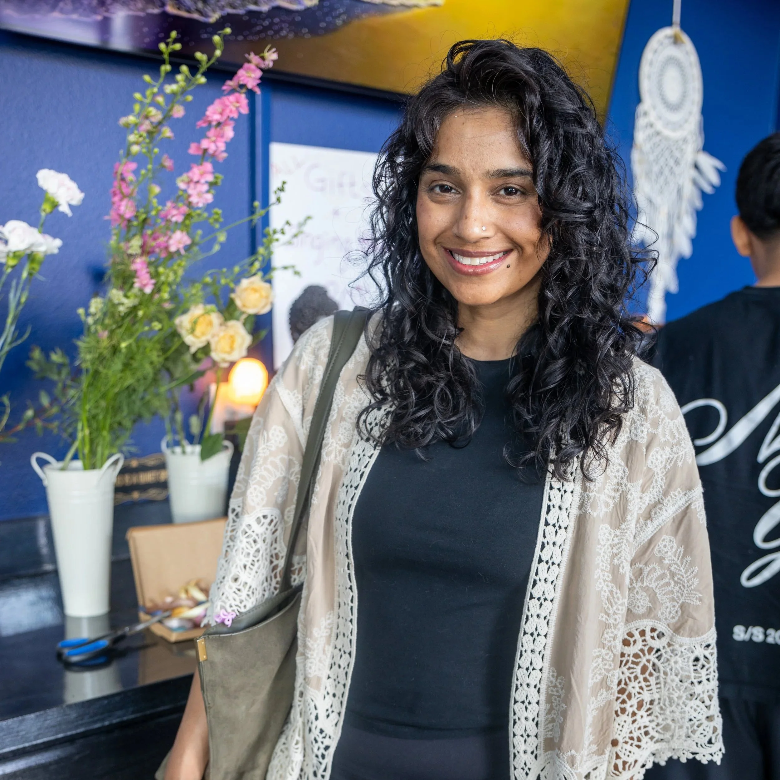A woman with black, curly hair smiling indoors, standing in front of a blue wall with various objects and flowers around her.