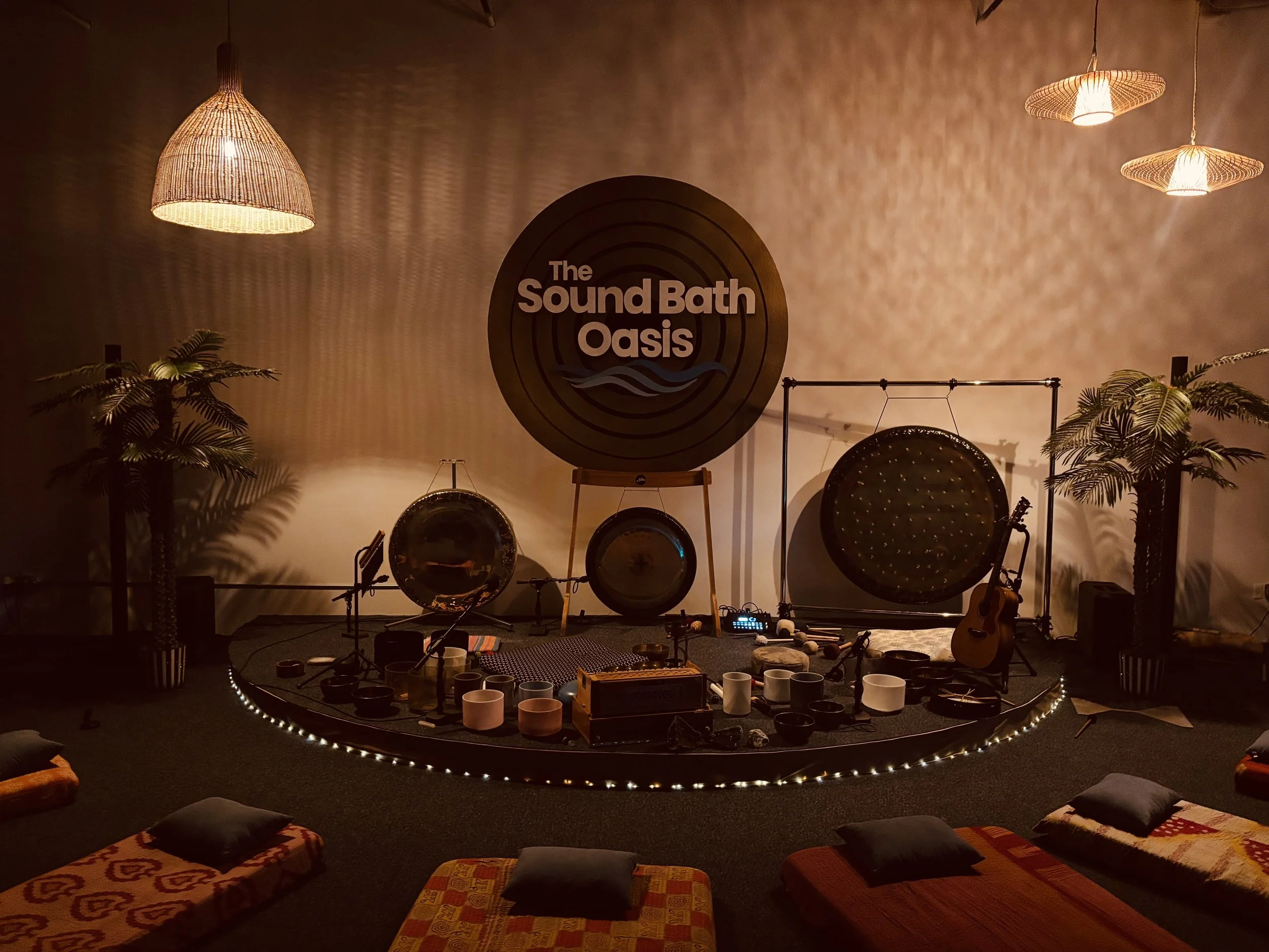 Music and meditation studio with drums, guitars, and singing bowls arranged in circle, surrounded by cushions and plants, warm lighting, and a sign that reads 'The Sound Bath Oasis' on the wall.