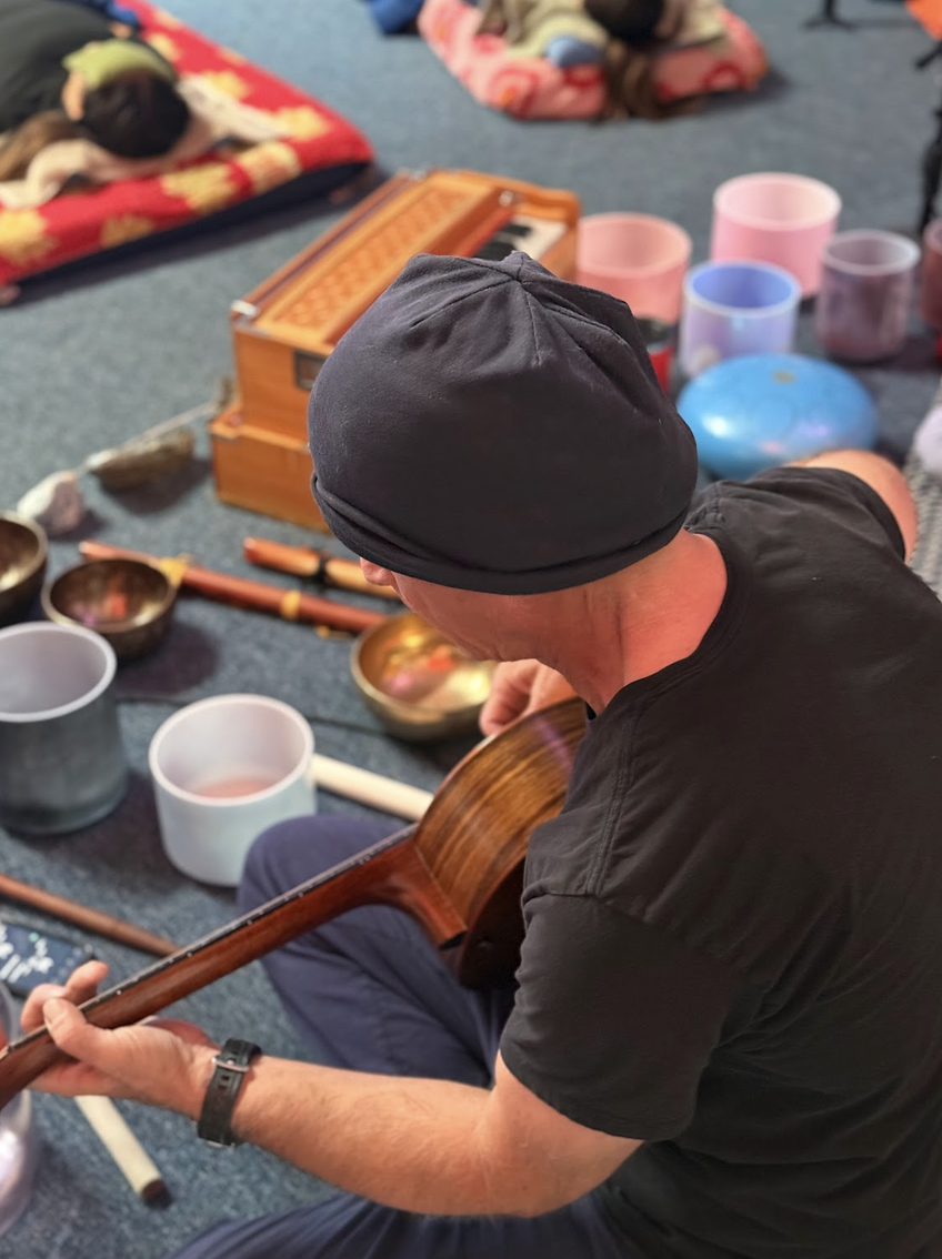 A man wearing a black beanie and shirt playing an acoustic guitar amidst singing bowls and percussion instruments on the floor, with people resting on cushions in the background.