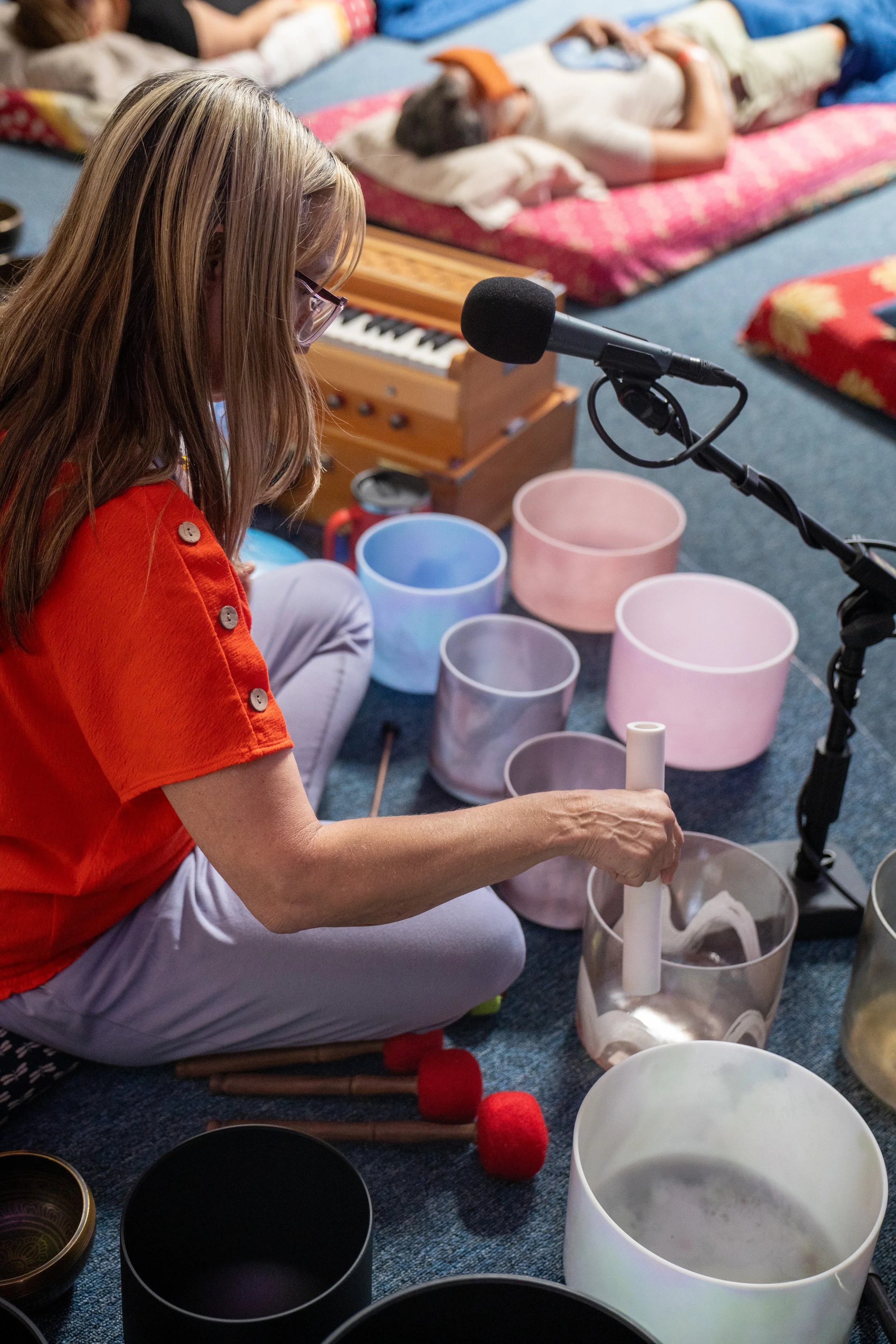 A woman wearing glasses and a red shirt is sitting cross-legged on the floor, playing a white handpan drum. She is surrounded by several other musical instruments, including steel tongue drums and a teak wooden keyboard. In the background, a child is