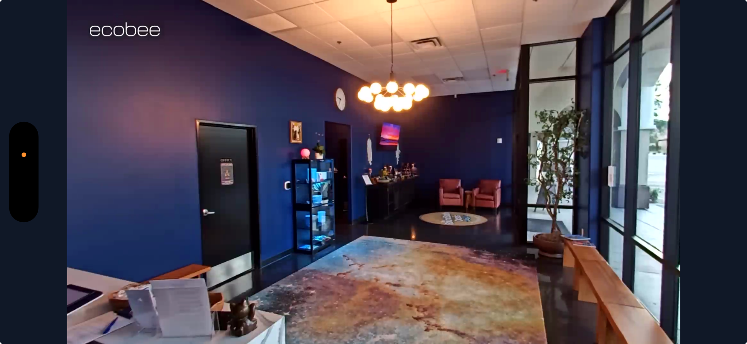 Interior of a modern office lobby with a dark blue accent wall, a colorful area rug, a sitting area with pink chairs, decorative lighting, and a glass entrance door.