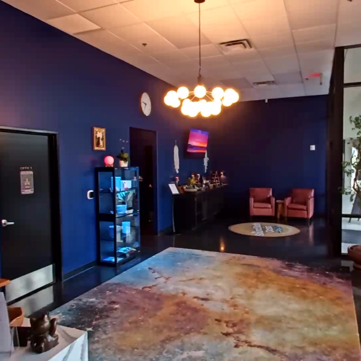 Lobby area with dark blue walls, a chandelier with multiple bulbs, pink chairs, a blue cabinet, a colorful painting, and a patterned rug.
