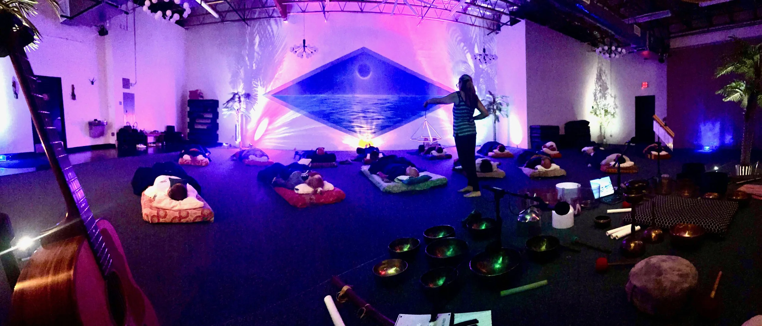 A group of people lying on cushions on the floor in a dimly lit room with purple lighting, while an instructor stands in front of them demonstrating breathing or meditation exercises. The background features a large mural of an ocean scene on the wal
