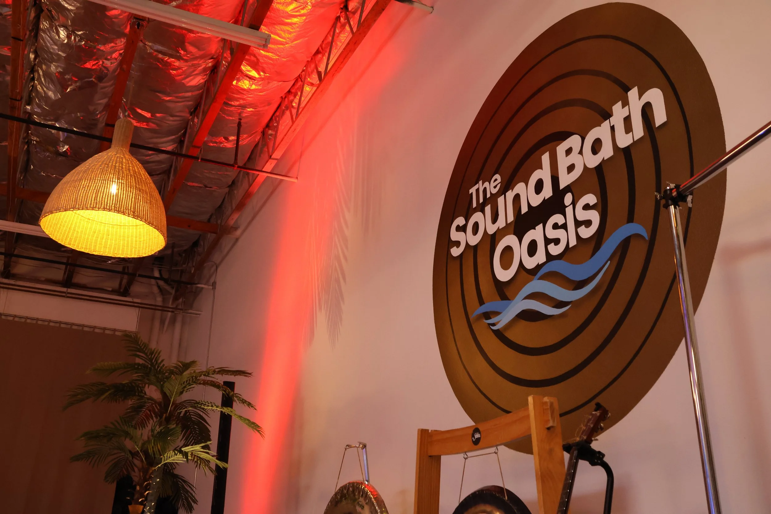 Interior of a space with exposed ceiling beams, silver insulation, a woven rattan pendant light, potted plant, and a wall display for The Sound Bath Oasis with a circular design and stylized waves.