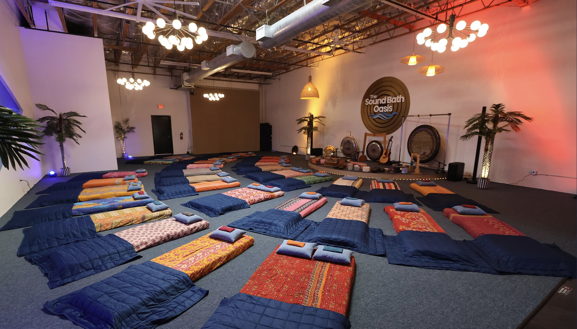 A meditation or yoga space with rows of colorful mats and cushions on a carpeted floor, a stage with musical instruments and a sign reading 'The Sound Bath Oasis,' and decorative plants under contemporary lighting fixtures in an industrial-style room