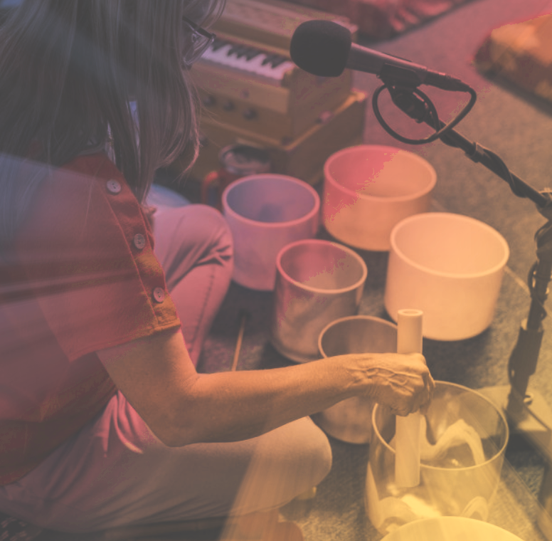 A woman playing crystal singing bowls and a wind gong, with a microphone nearby, in a cozy indoor setting.