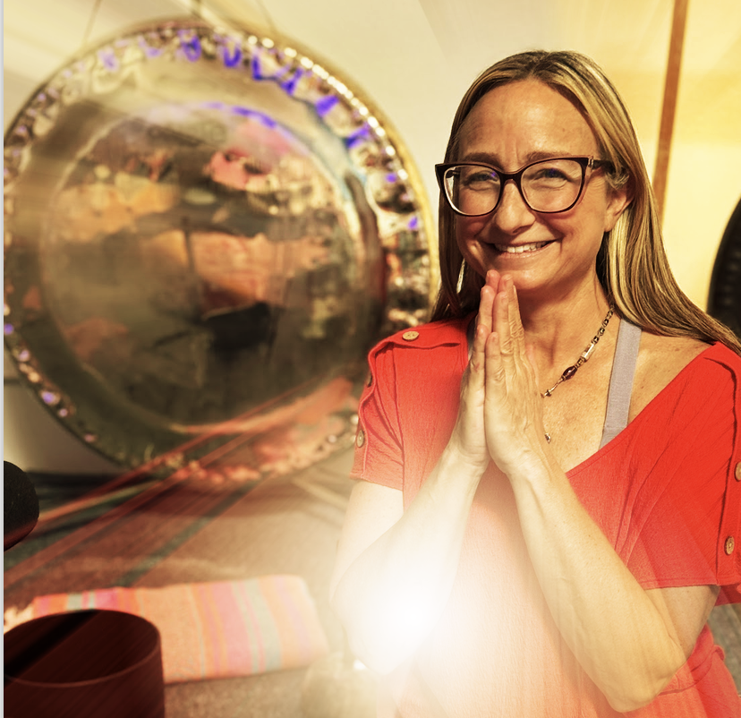 A woman with glasses, smiling and holding her hands together near her face indoors, with a large reflective gong in the background.