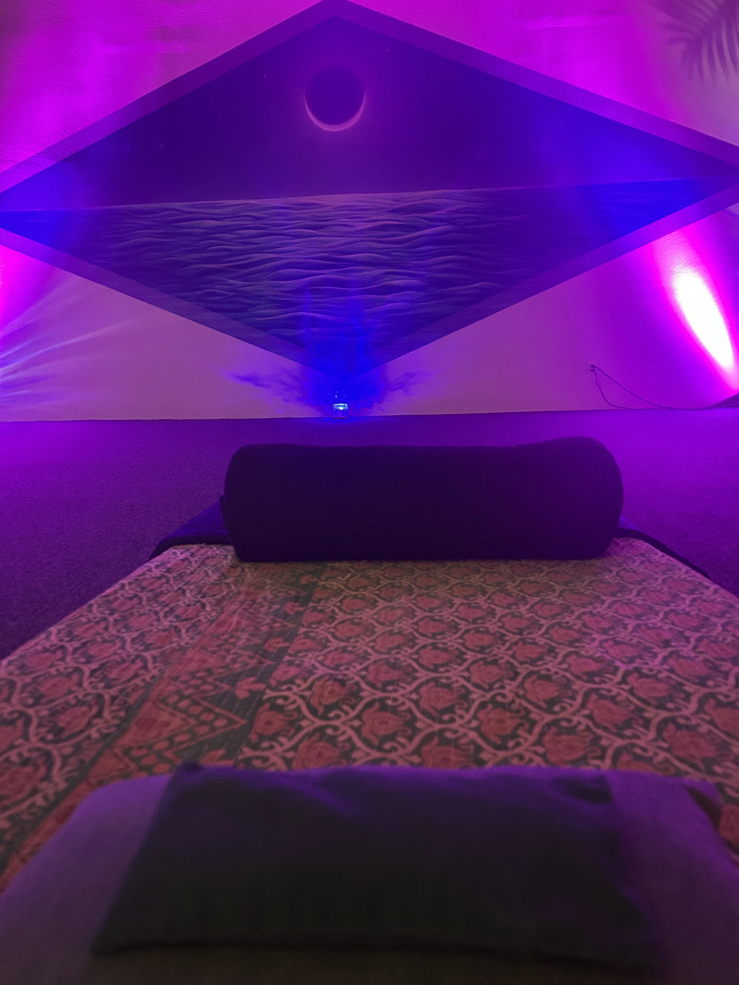 View from a bed with a patterned bedspread, looking toward a wall with purple and pink lighting. A black pillow is on the bed, and a projection on the wall depicts a moon over water with a blue and purple color scheme.