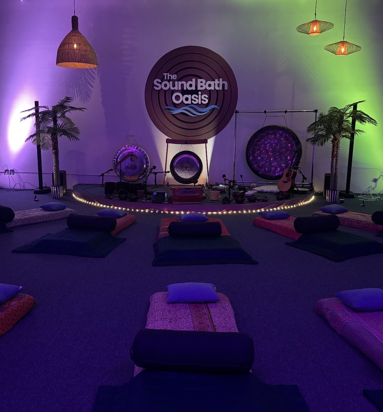Indoor stage decorated for a sound bath session, with a sign reading 'The Sound Bath Oasis'. The stage features meditation cushions, a guitar, gongs, and various instruments, with palm plants and colorful ambient lighting.