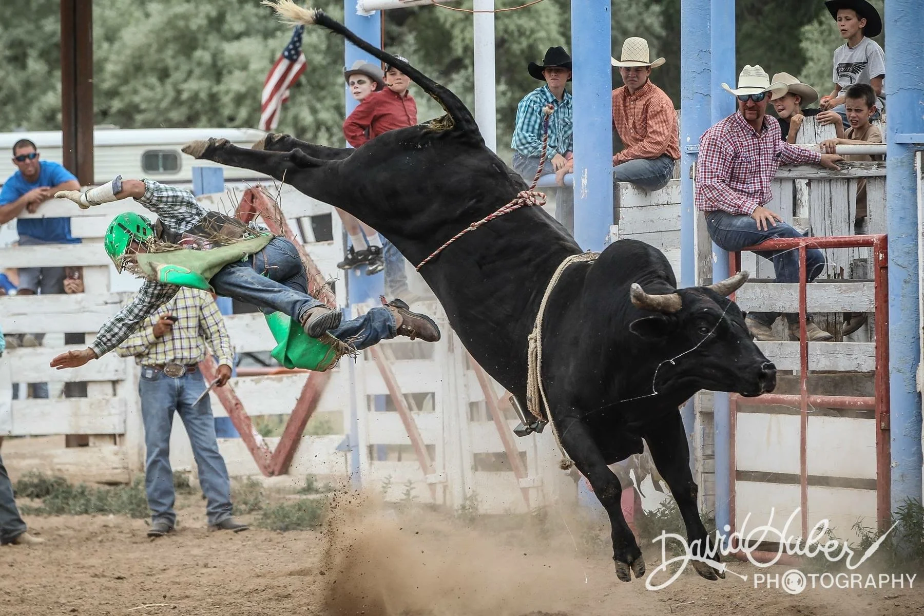 A rodeo scene with a person on a bucking black bull, wearing a green helmet and plaid shirt, falling off as the bull leaps into the air. Spectators watch from behind a white and blue fence, with some sitting on raised platforms and others standing ne