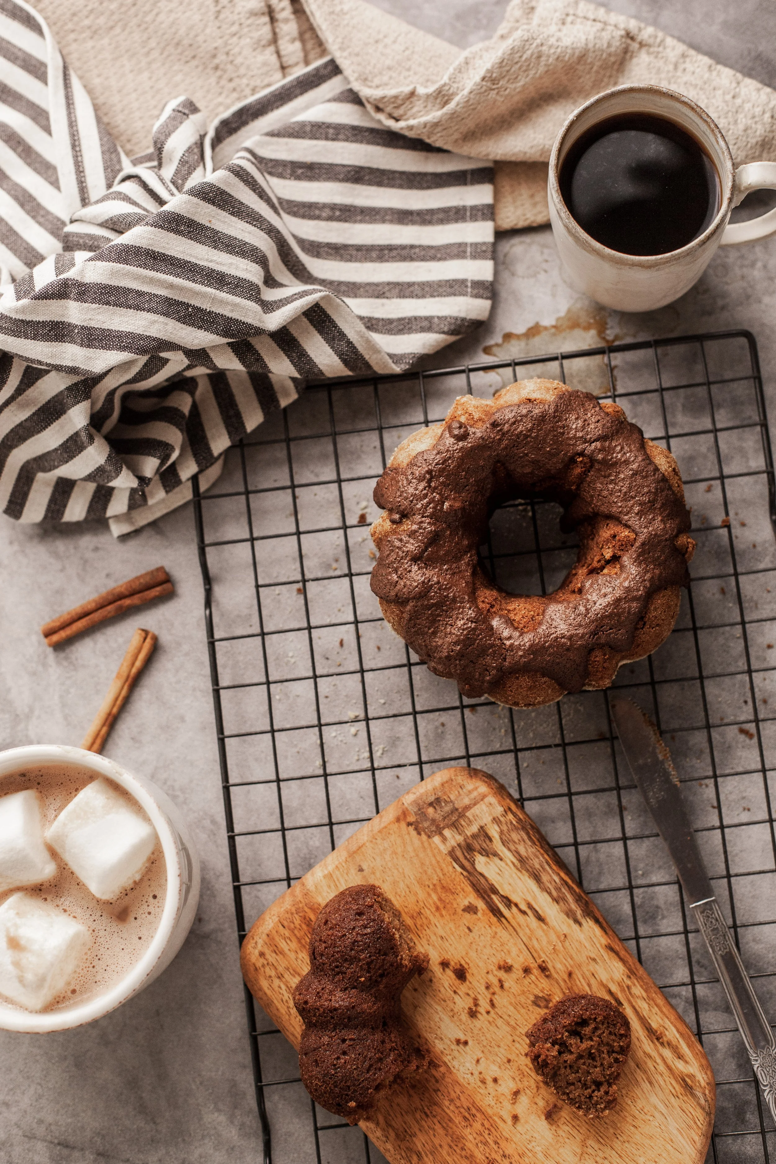 Chocolate Bundt cake on a cooling rack, with a knife, a wooden cutting board, a mug of coffee with marshmallows, and cinnamon sticks on a gray surface, topped with a striped cloth and a beige towel.