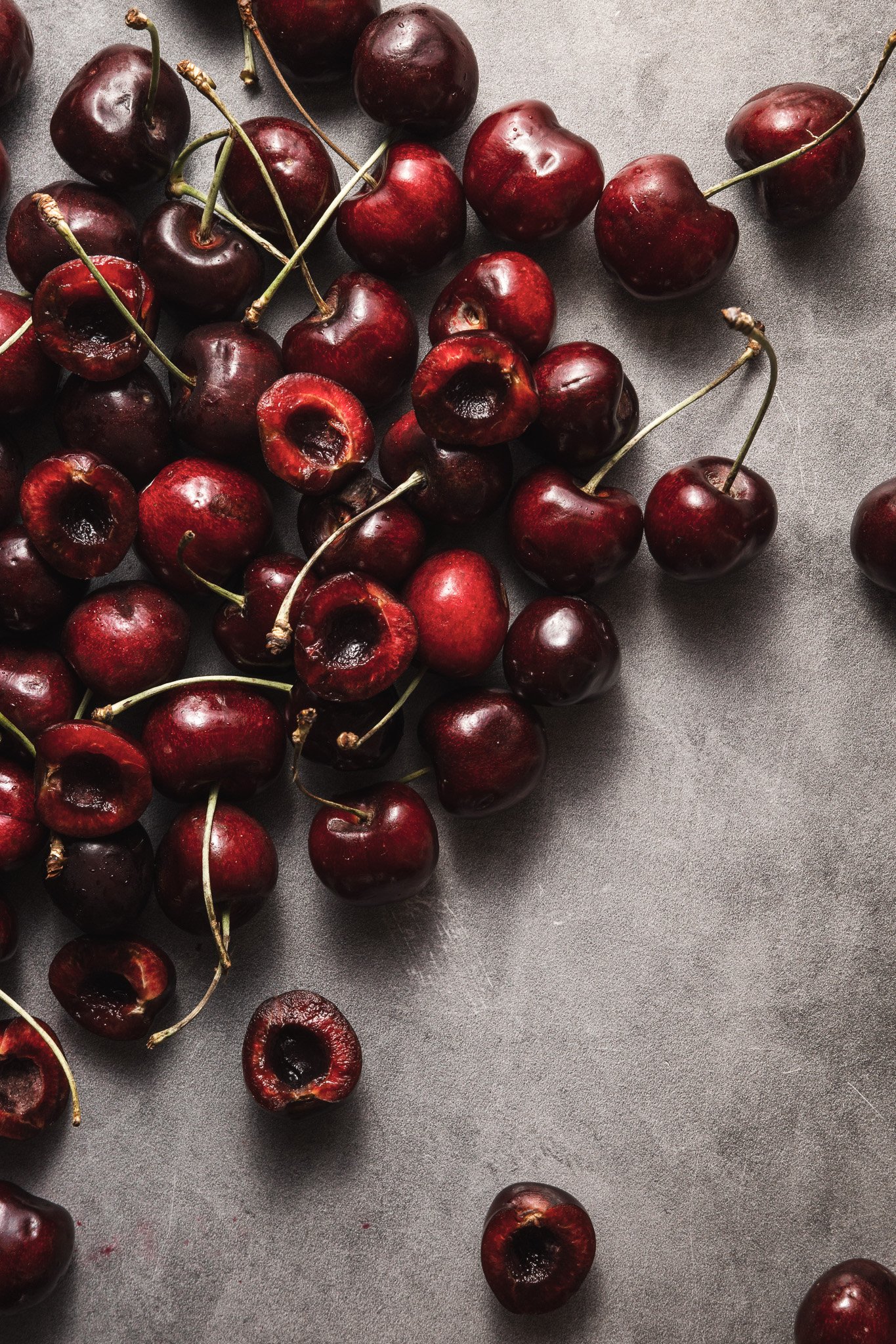 A collection of fresh dark red cherries with green stems, some with pits exposed, scattered on a gray surface.