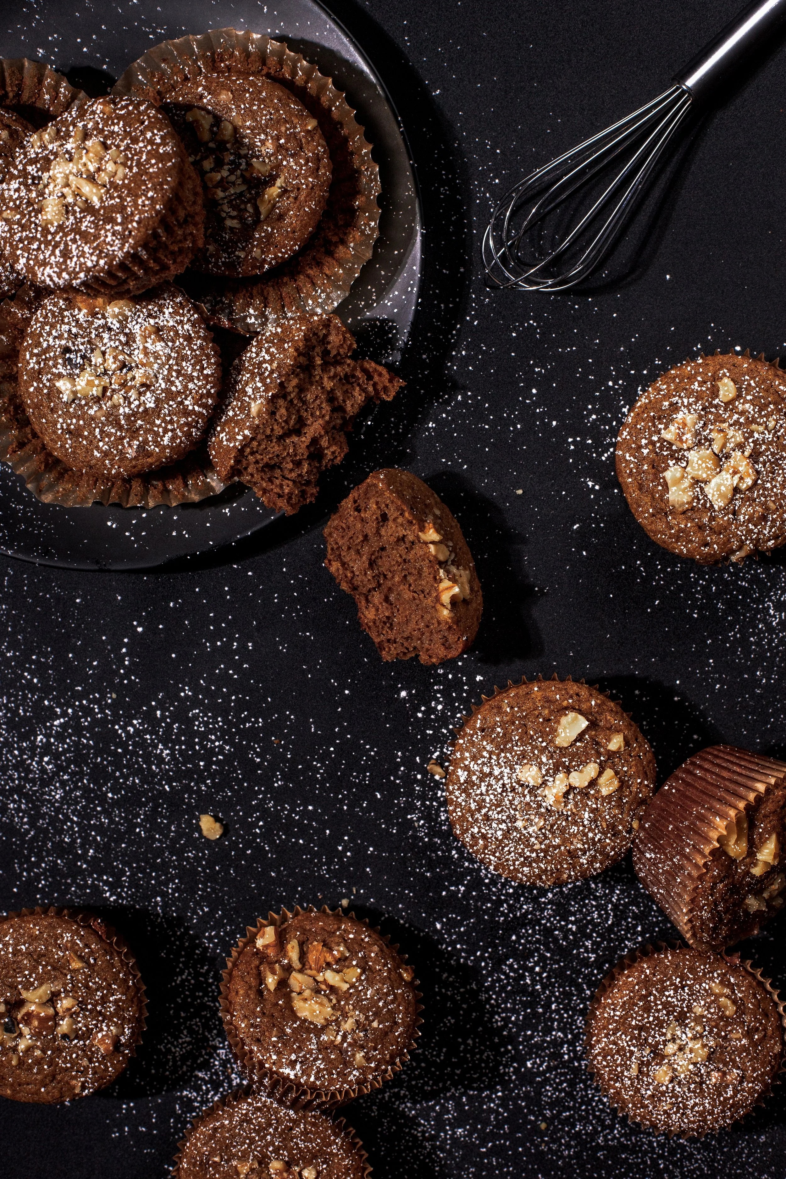 Chocolate muffins topped with chopped nuts, sprinkled with powdered sugar, on a black surface with a metal whisk nearby.