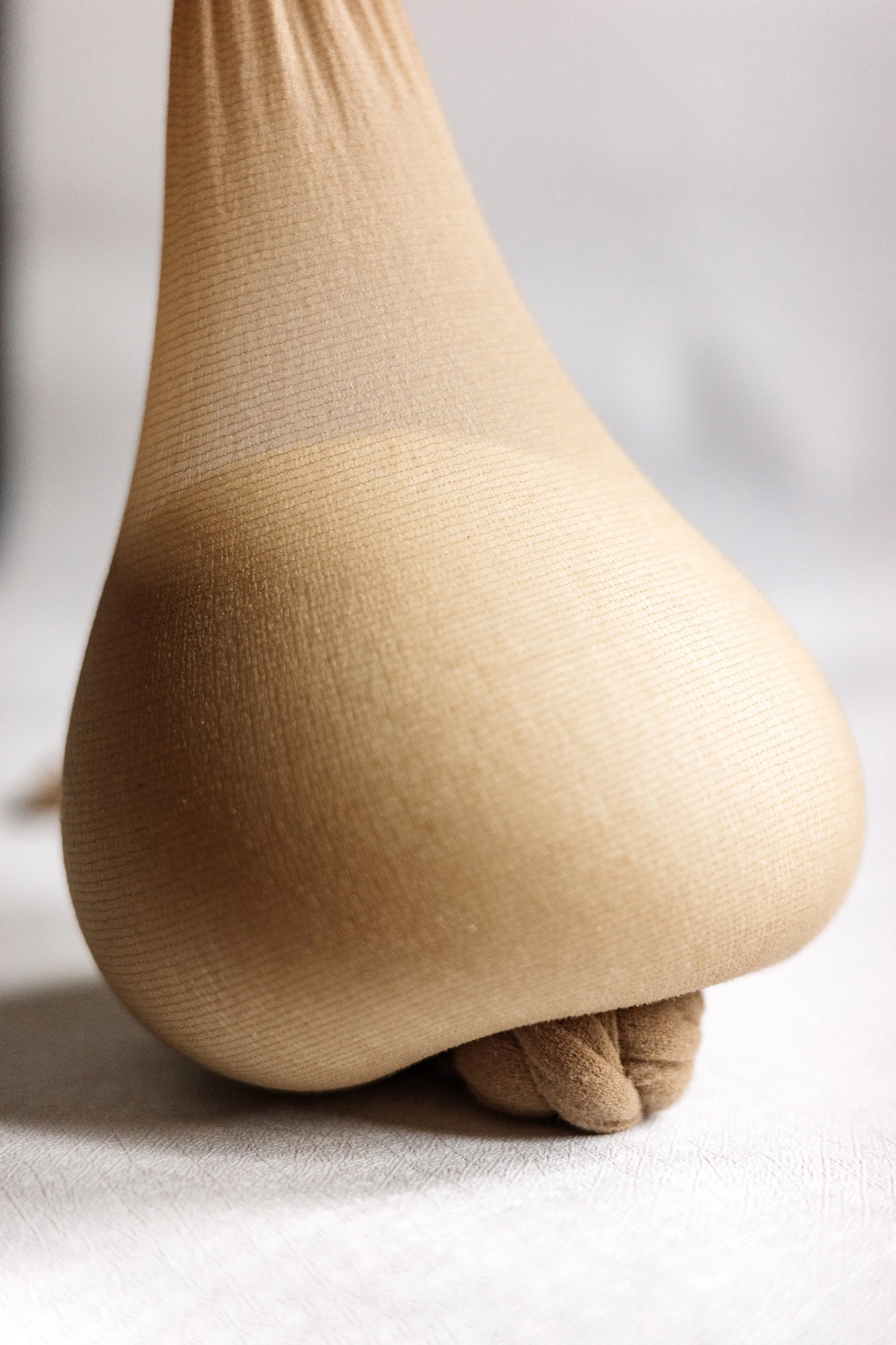 Close-up of a mannequin's leg wearing nude-colored tights and brown shoes, set against a plain background.
