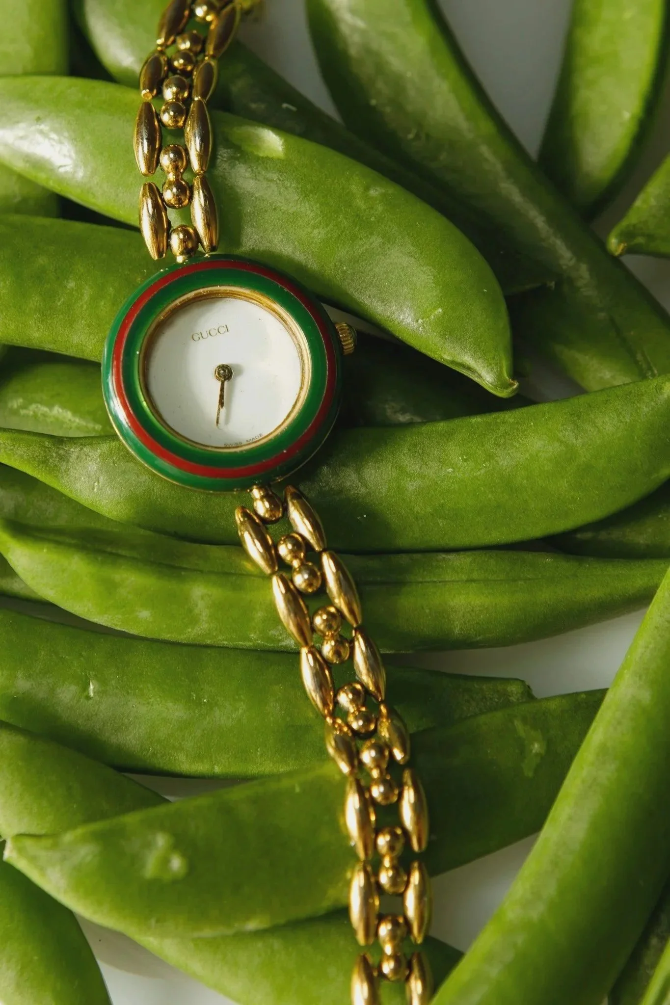 A Gucci wristwatch with a gold link bracelet and a white dial, surrounded by green sugar snap pea pods.