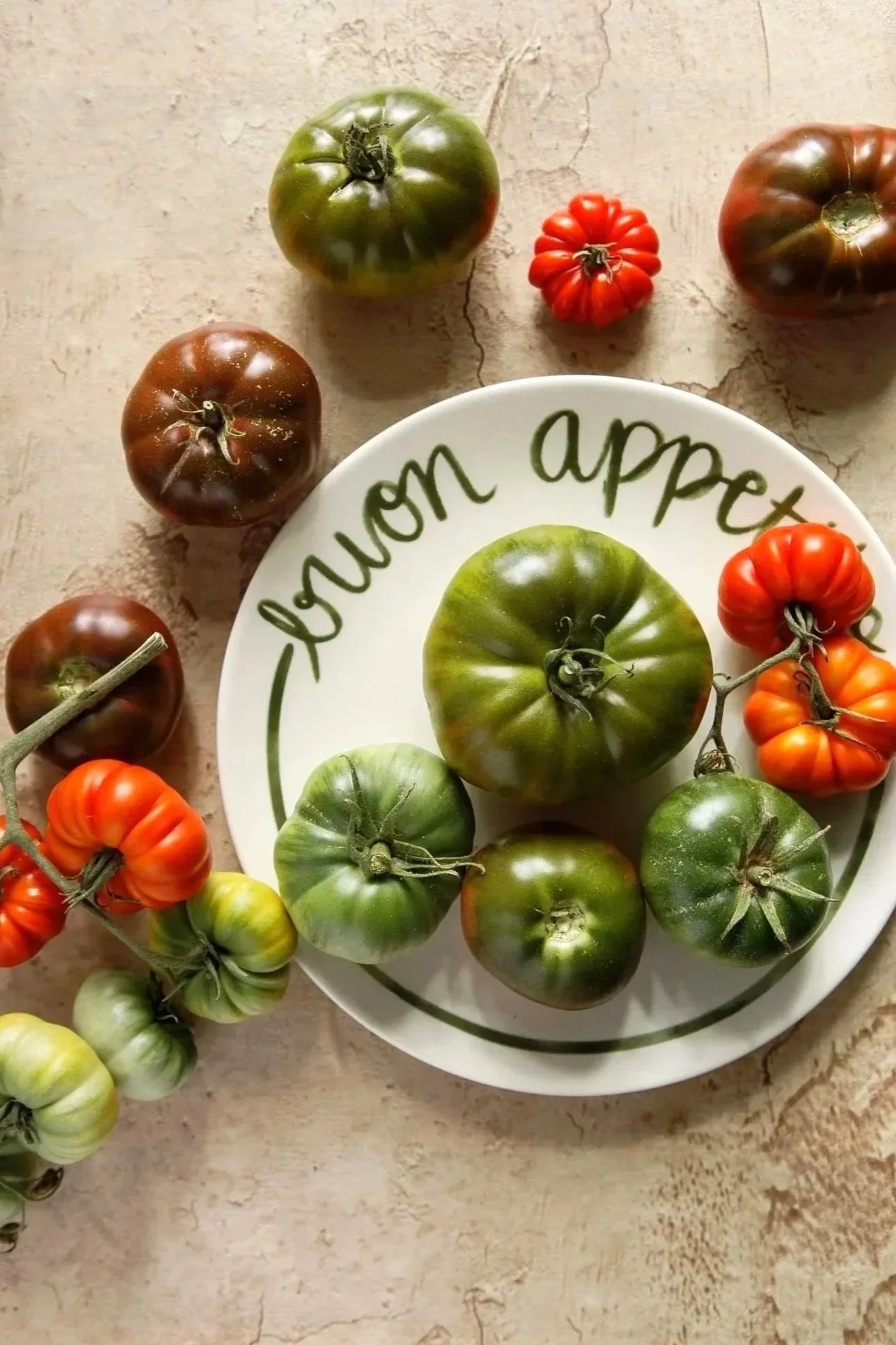 A white plate with the words "buon appetito" written on it, surrounded by various heirloom tomatoes in different colors and sizes, on a textured surface.