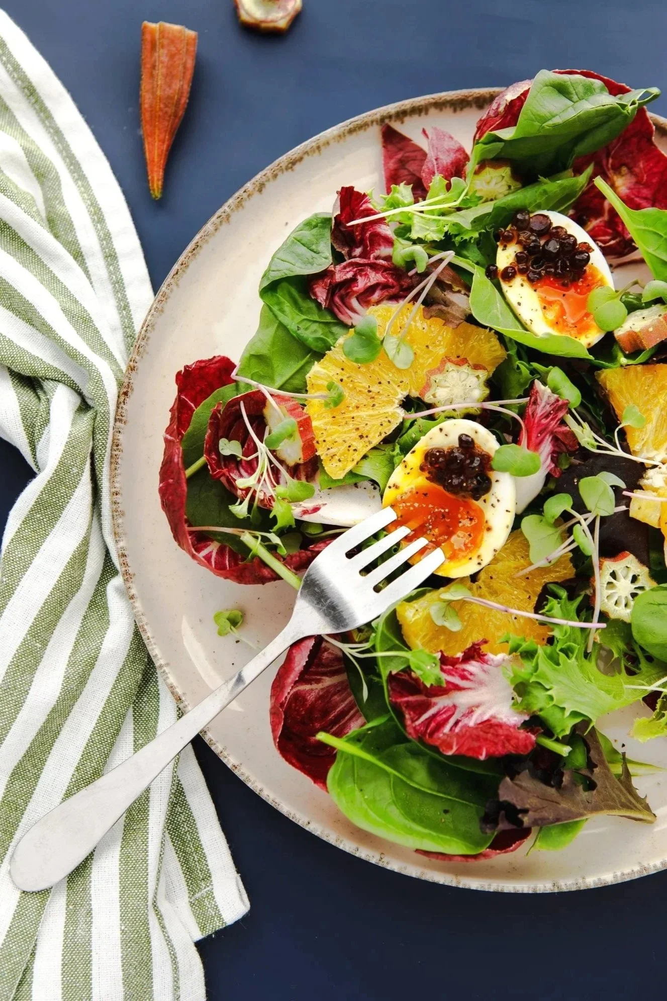 Radicchio salad with soft-boiled eggs, oranges, mixed greens, radicchio, and okra on a blue background and with a striped towel. 