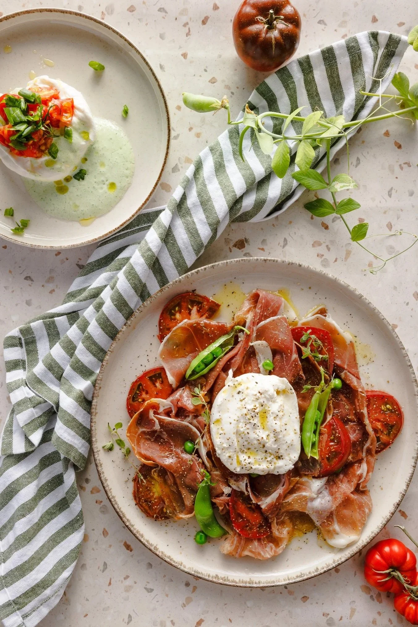 Plate of prosciutto, sugar snap peas and pods, fresh thyme sprigs, and burrata surrounded by tomatoes with a striped towel on a terrazzo background. 