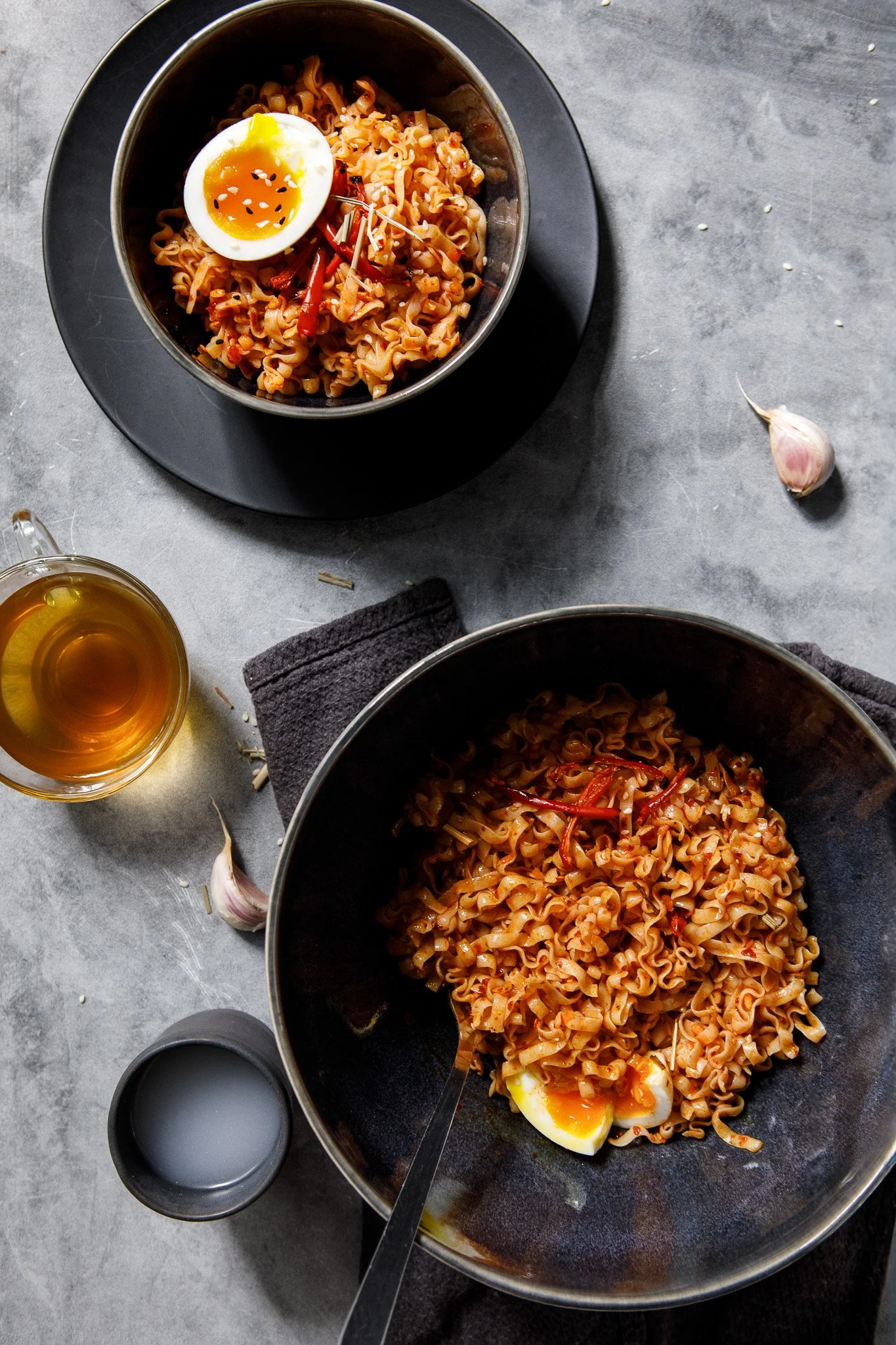 Tight vertical flat-lay showing noodle bowls, soft-boiled egg half, rice bowl, small dark cup, tea glass, garlic clove, and grey linen napkins.