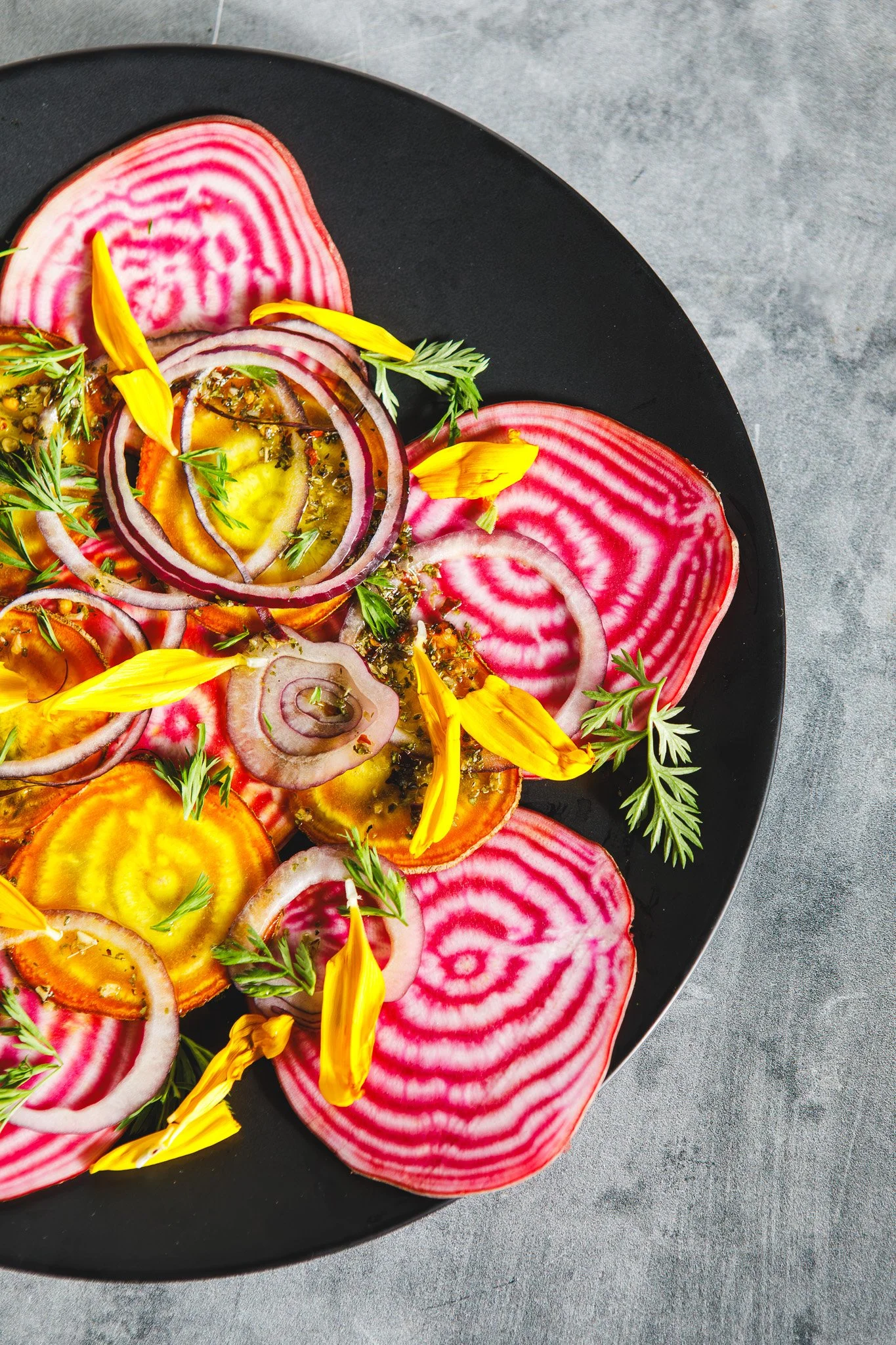 A plate of sliced beets with a pink and white spiral pattern, garnished with yellow flower petals and green herbs, on a gray surface.