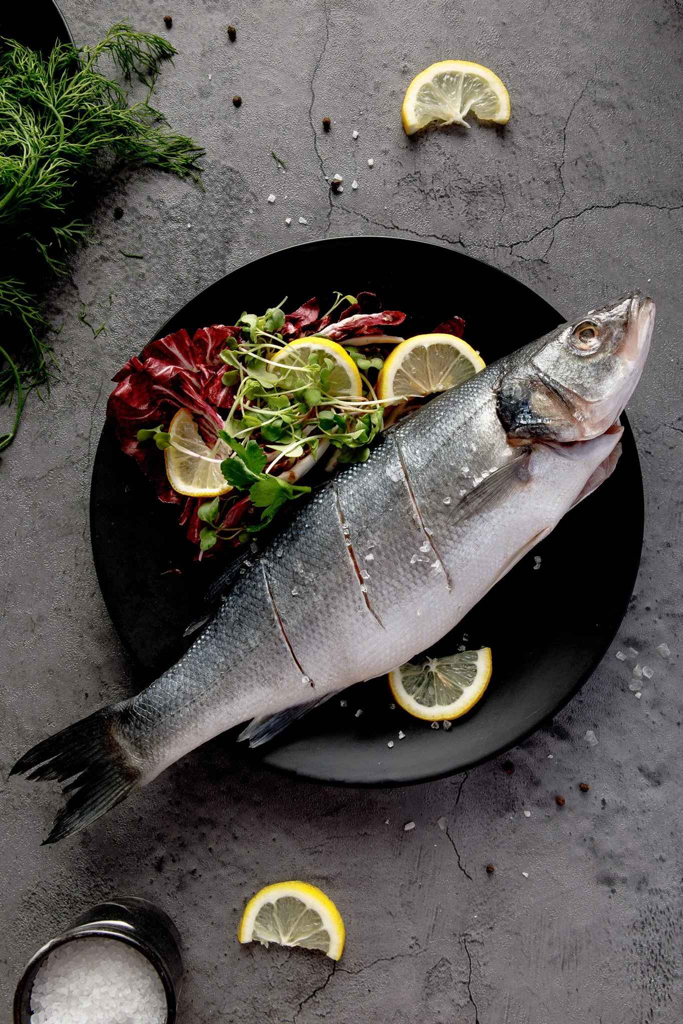 Whole fish on a black plate garnished with lemon slices, microgreens, and radicchio, on a textured gray surface surrounded by lemon slices, salt, and scattered peppercorns.