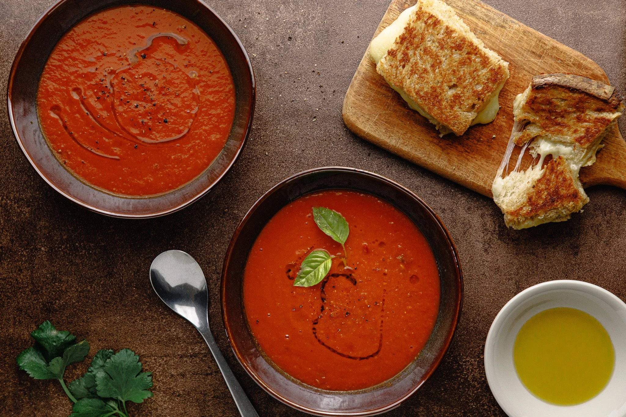 Wide top-down overhead (flat lay) showing bowls of tomato soup inspired by an NYT Cooking recipe, a wooden board with grilled cheese, herbs, a spoon, and an oil ramekin balanced across the frame.