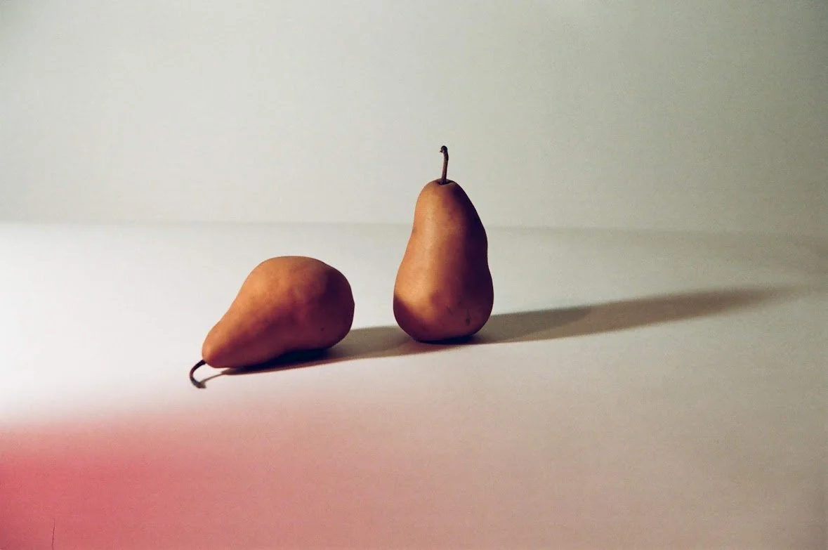 A ripe pear and an unripe pear placed on a white surface with a light background, casting distinct shadows.