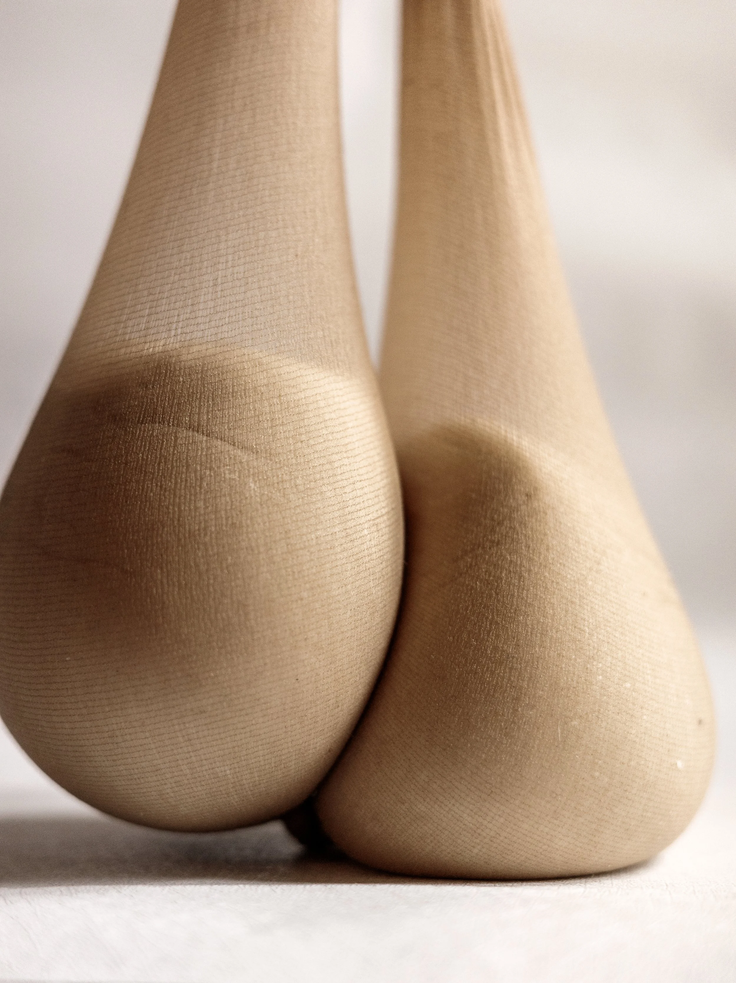 Close-up of a pair of legs wearing sheer beige stockings or tights, resting on a white surface, with a plain background.