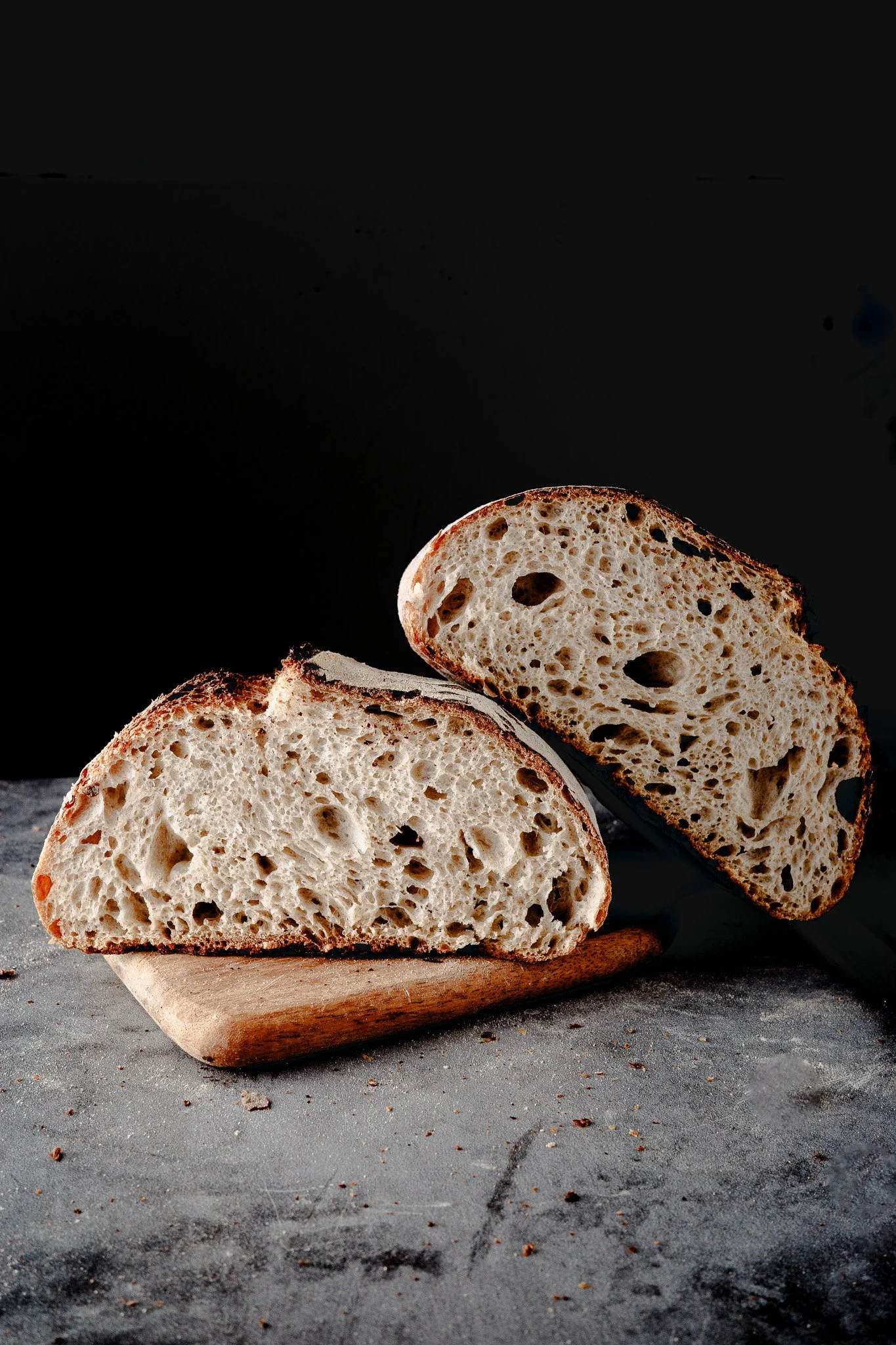 Slices of rustic sourdough bread with a crusty exterior and airy interior arranged on a wooden cutting board on a gray textured surface.