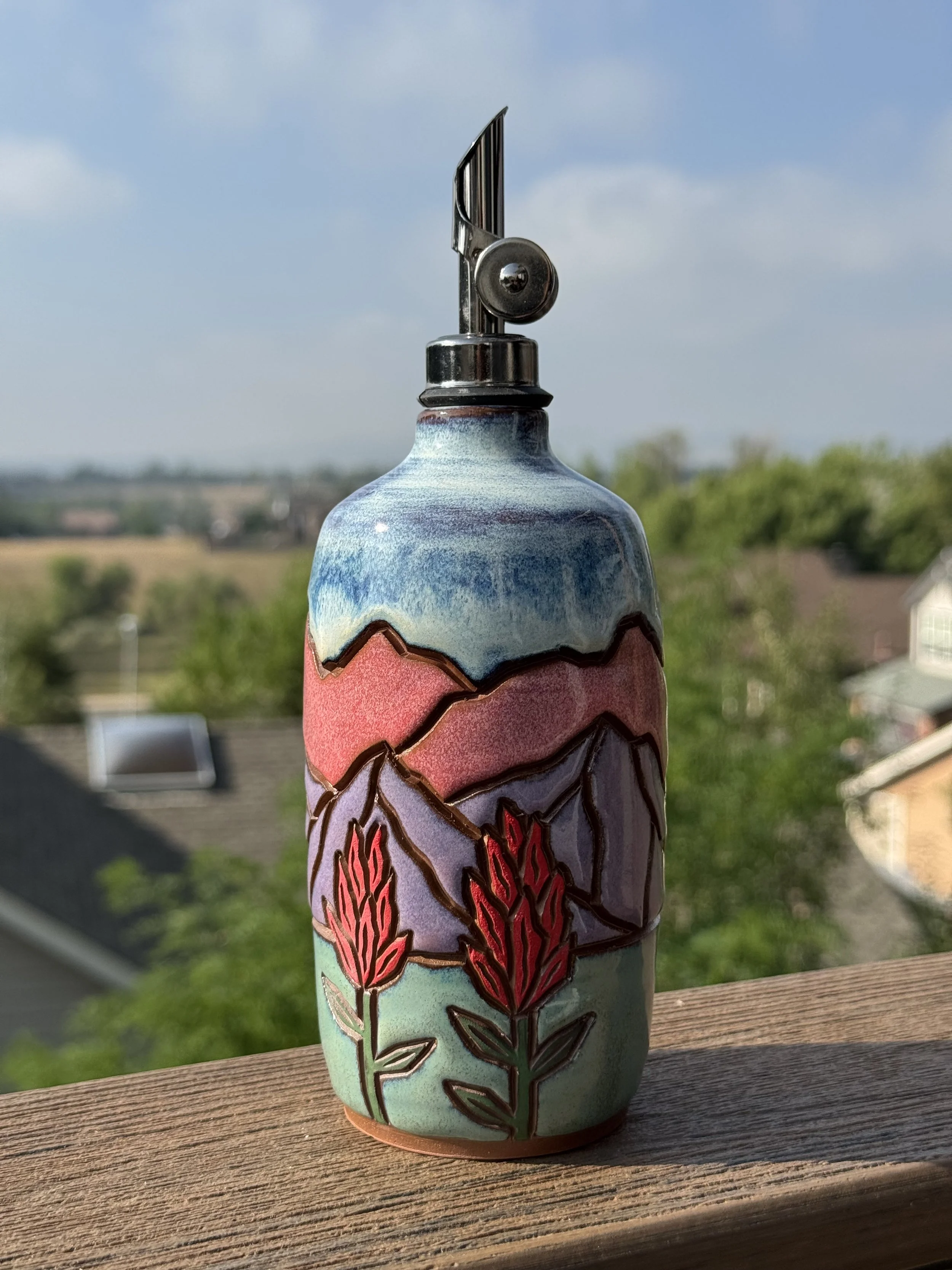 A colorful ceramic vase with a mountain and floral design, placed on a wooden surface outdoors with a blurred background of trees.