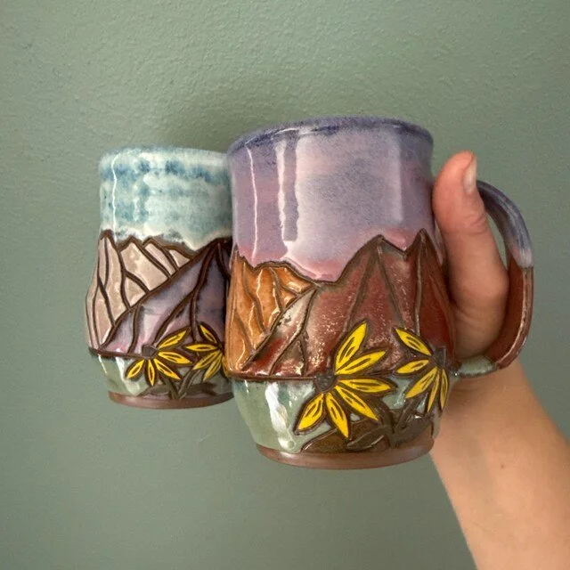 Two ceramic coffee mugs featuring mountain and leaf designs, held in a person's hand.