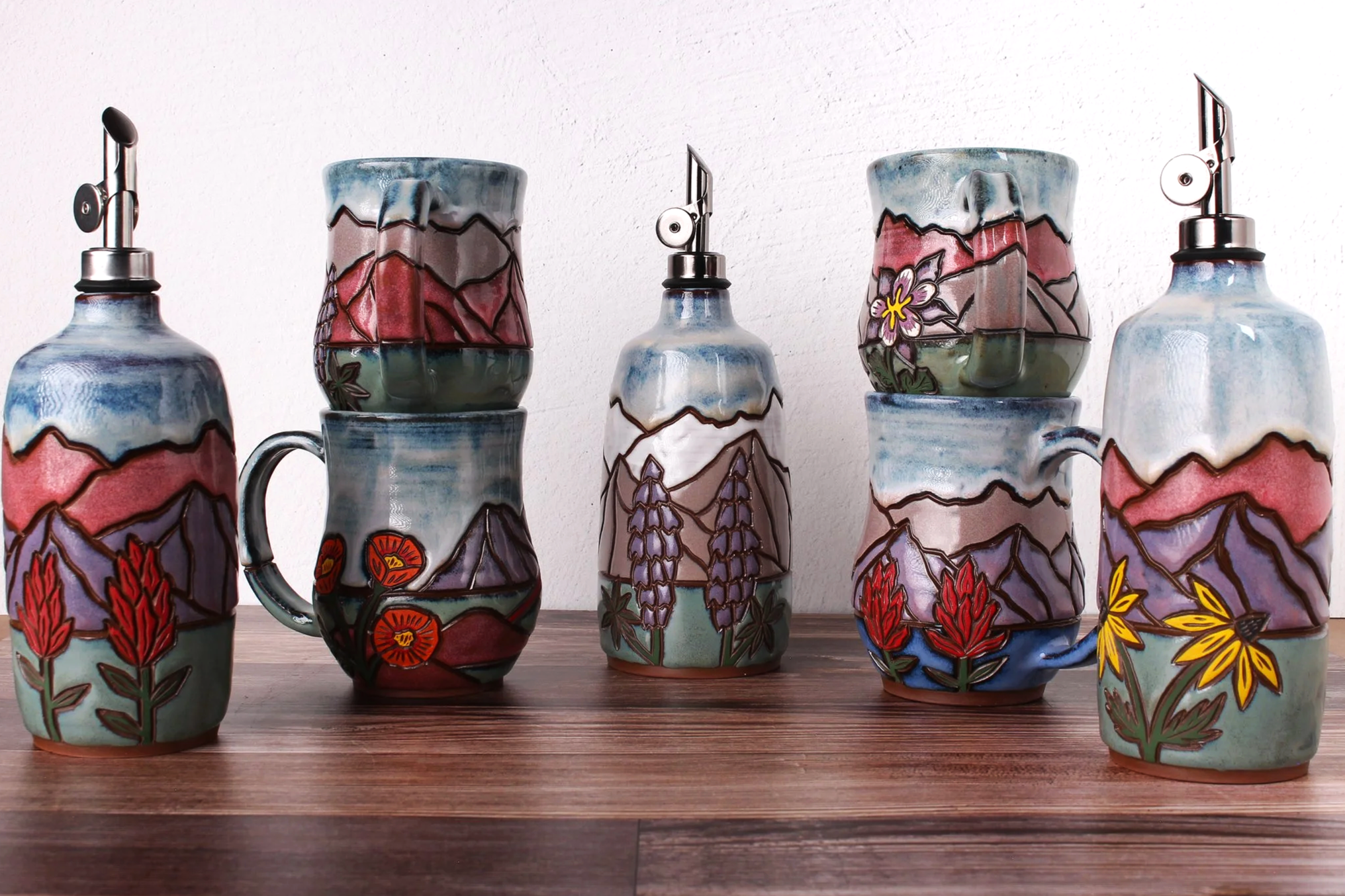 Collection of ceramic mugs with colorful mountain and floral designs, arranged on a wooden surface against a white wall.